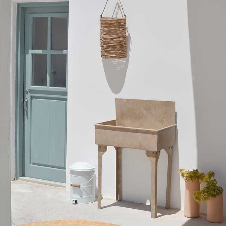 Fioriera beige in pietra con gambe davanti a muro bianco, porta azzurra e cesta di vimini appesa