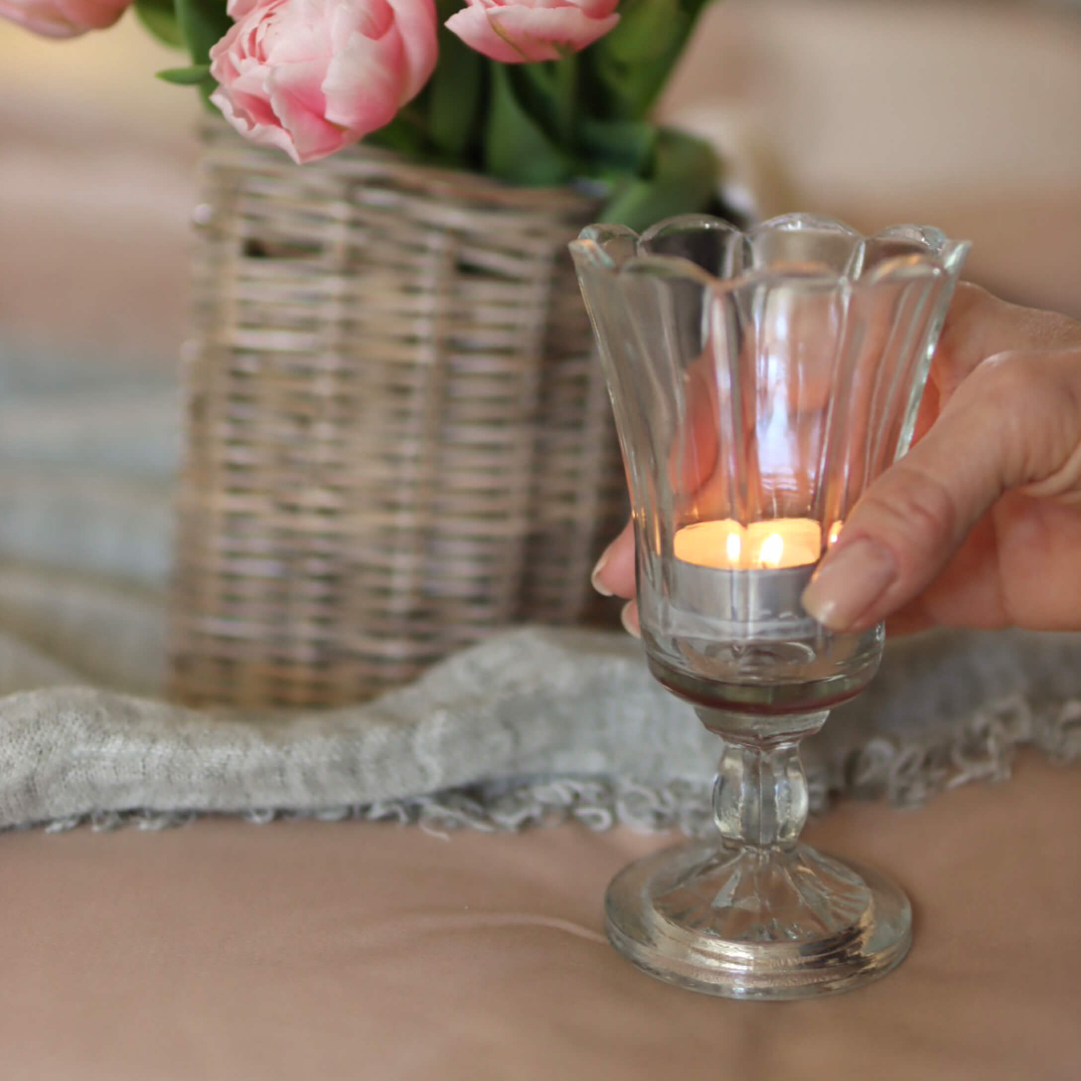 Portacandela in vetro su piede con candela accesa, vaso con fiori rosa e cesto di vimini sullo sfondo