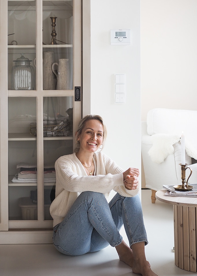 Donna sorridente con maglione bianco e jeans blu seduta a terra in un interno luminoso e accogliente