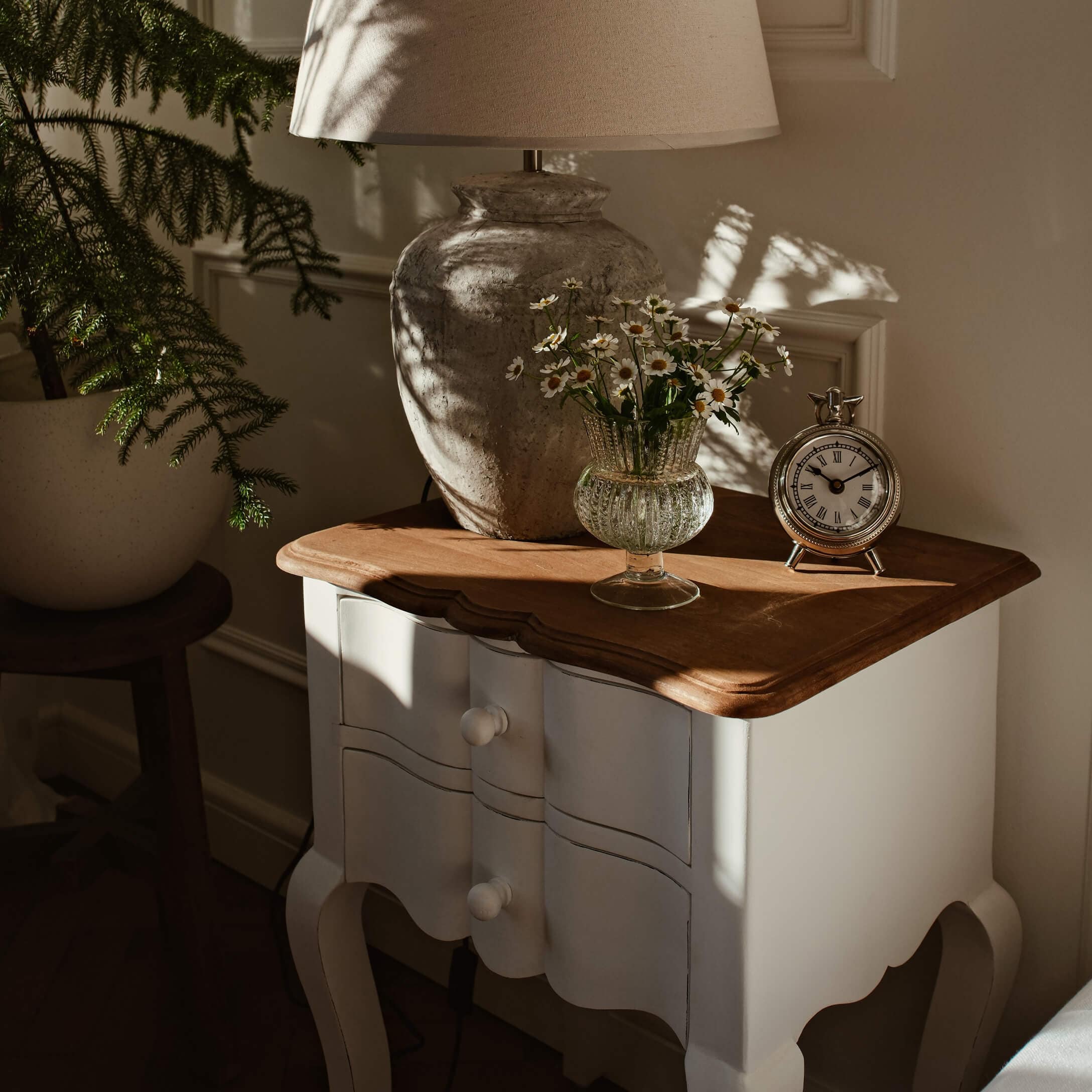 Comodino bianco in legno con piano marrone, lampada, vaso di vetro con fiori e orologio argento