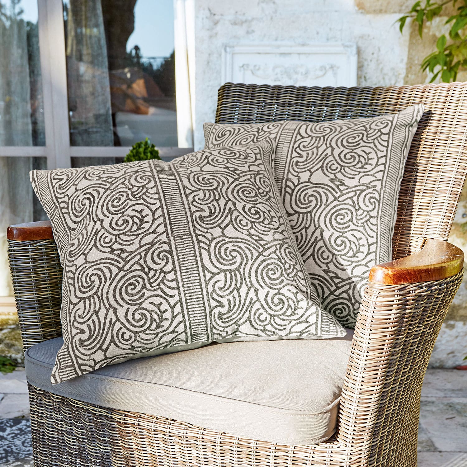 Coppia di copricuscini Ensis Due cuscini decorativi in cotone beige con motivo marrone su poltrona in vimini su terrazza