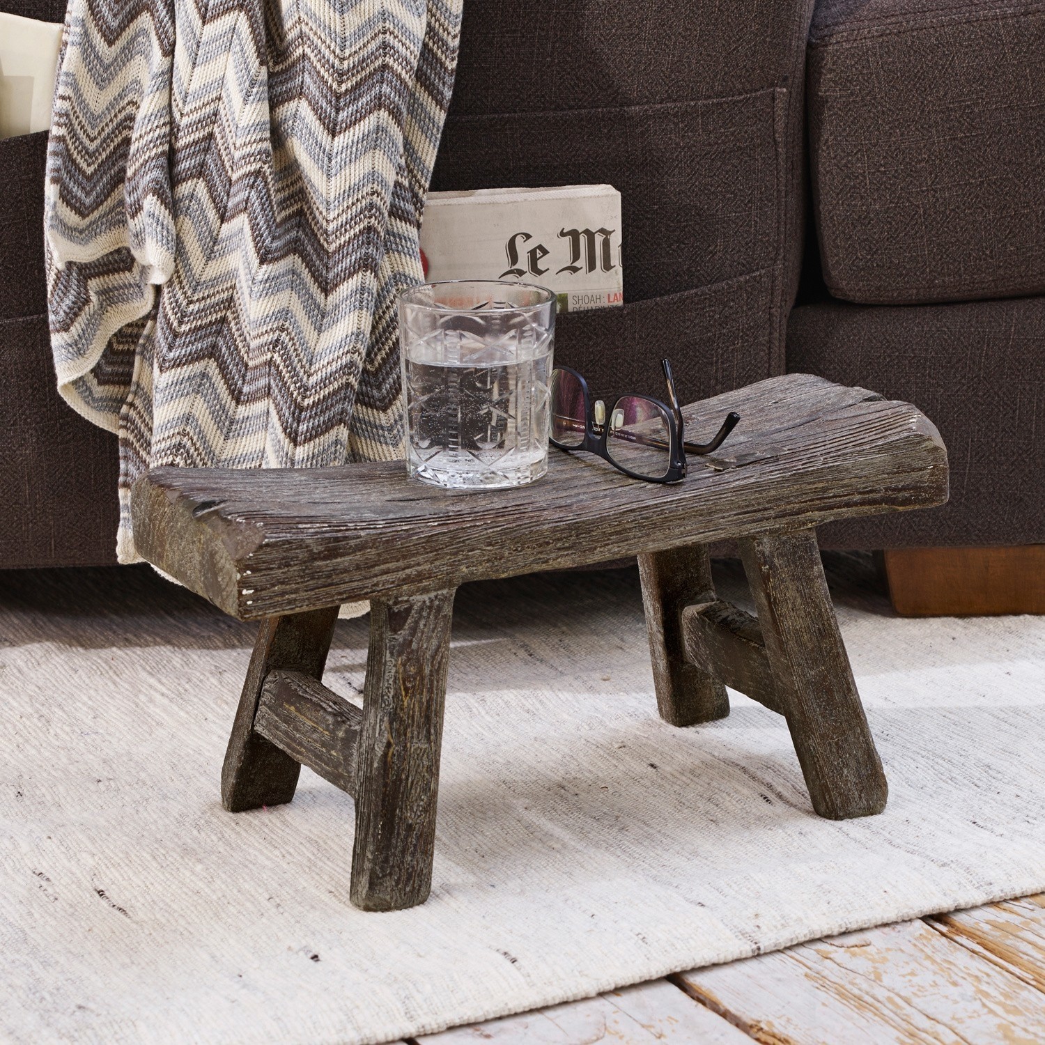 Sgabello Cadoc Tavolino rustico basso in legno scuro su tappeto chiaro accanto a divano con coperta a zigzag