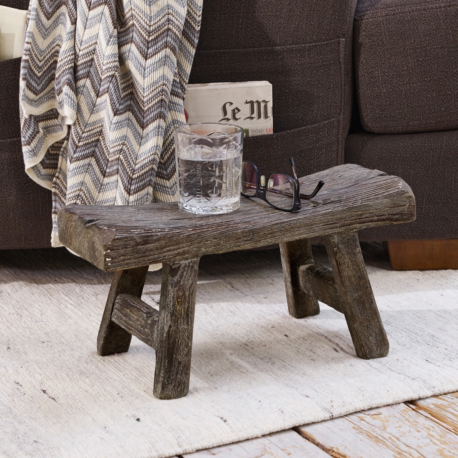 Sgabello Cadoc Tavolino rustico basso in legno scuro su tappeto chiaro accanto a divano con coperta a zigzag