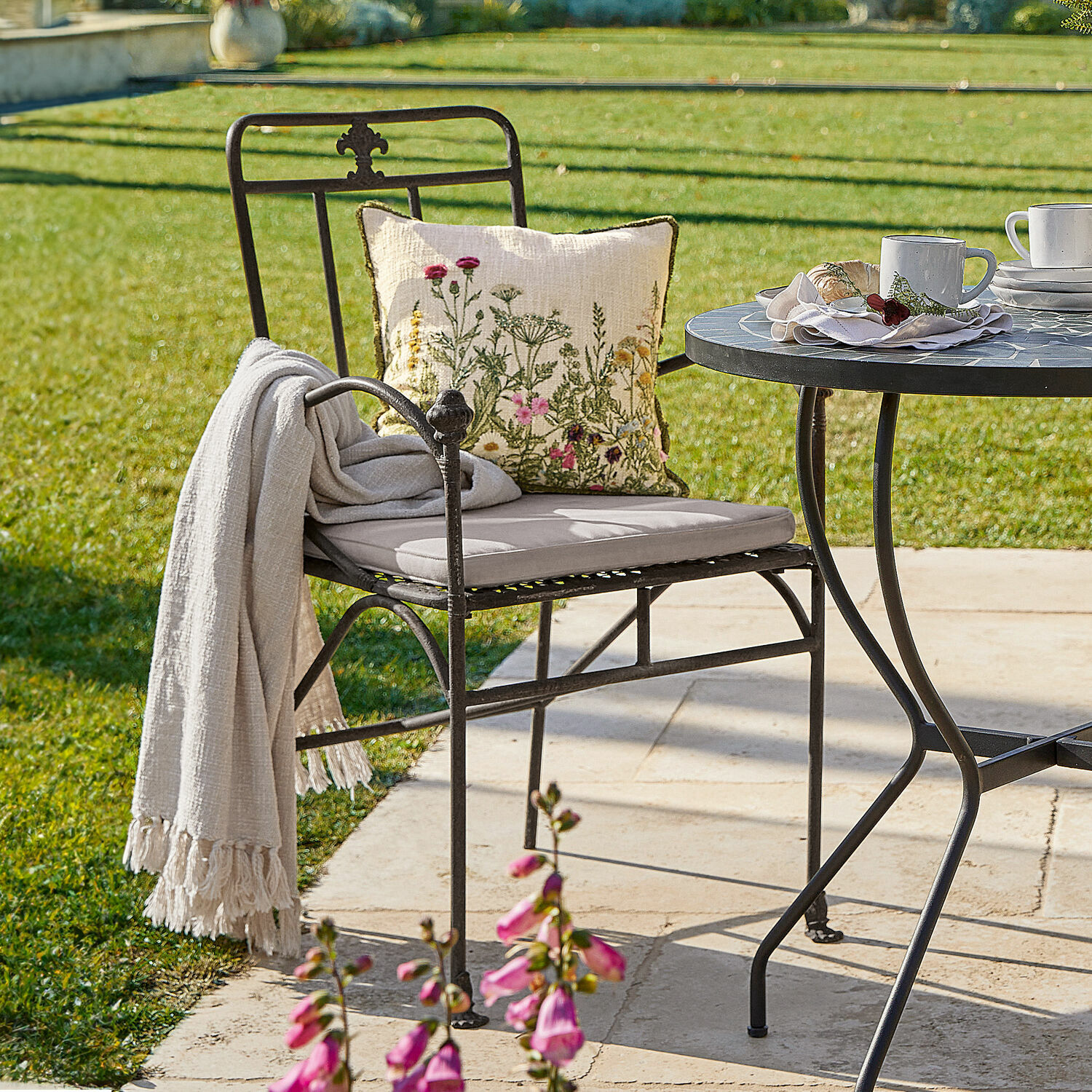 Sedia Maidis Sedia da giardino in metallo nero con cuscino chiaro e cuscino decorativo floreale su terrazza soleggiata