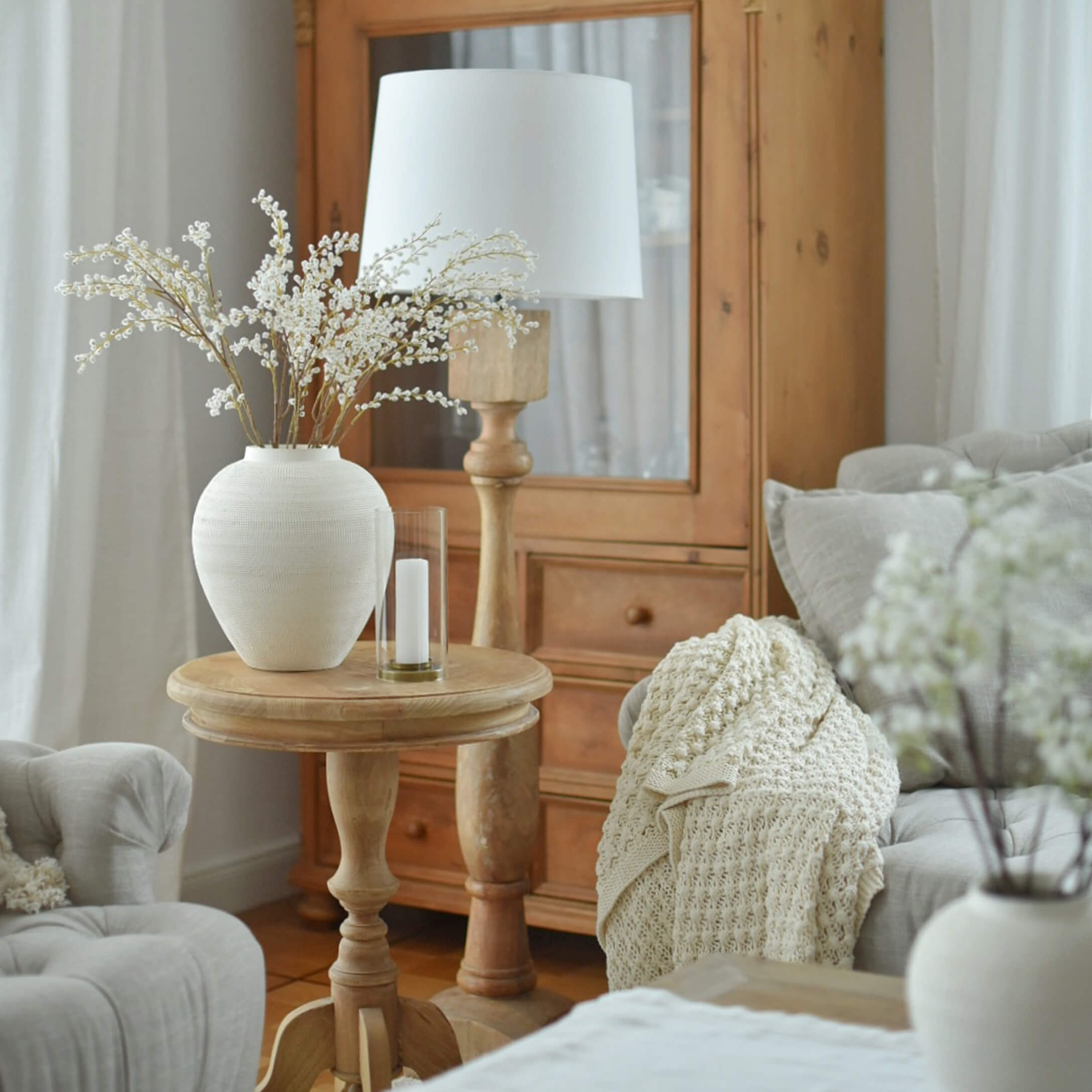 Tavolino in legno chiaro con vaso in ceramica bianca e lampada in soggiorno accogliente con coperta beige