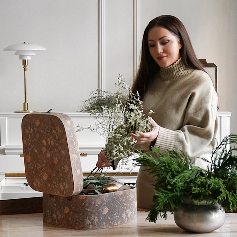 Donna con maglione beige che tiene rami verdi accanto a scatola decorativa e pianta in vaso argento