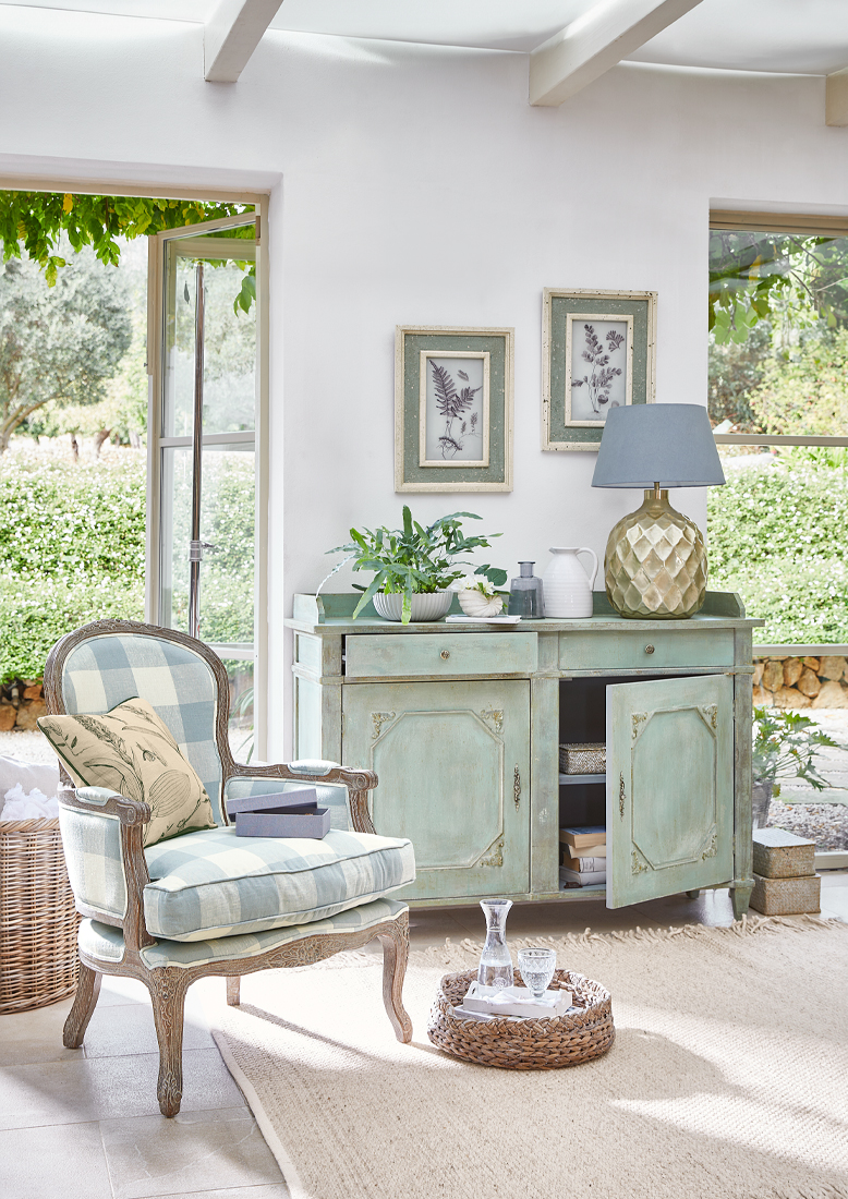 Credenza vintage in legno verde menta con lampada decorativa e piante in soggiorno luminoso con poltrona a quadri