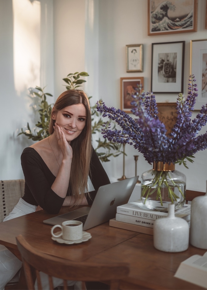 Donna seduta a tavolo in legno con laptop e vaso di vetro con fiori viola in interno luminoso