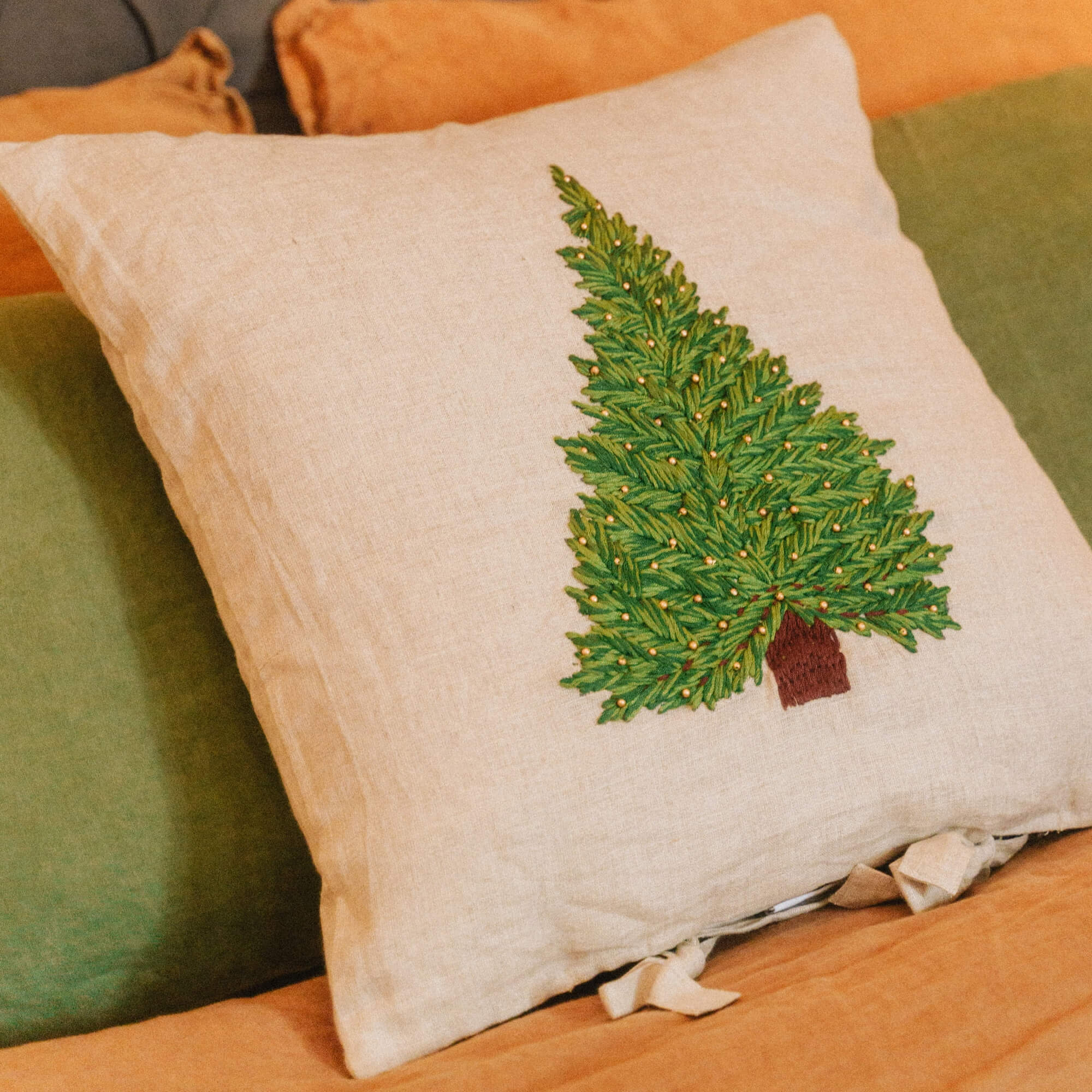Cuscino in lino beige con ricamo di albero di Natale verde e dettagli dorati su biancheria da letto arancione