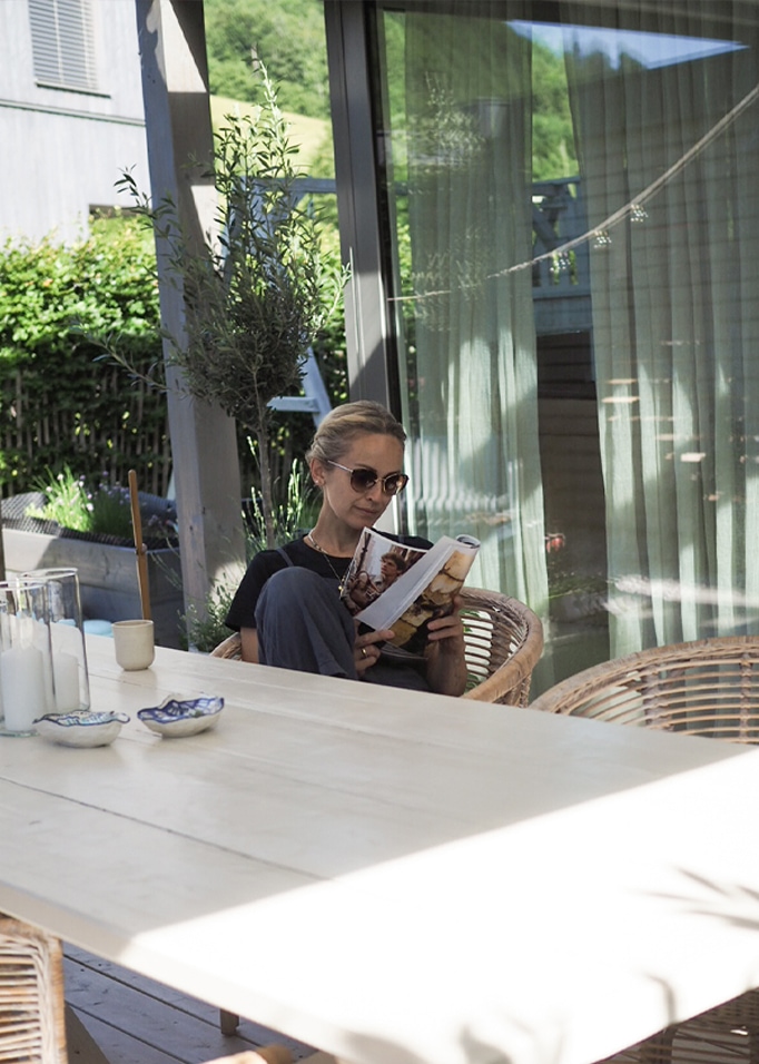 Donna che legge un libro su terrazza con poltrona in vimini e tavolo in legno chiaro