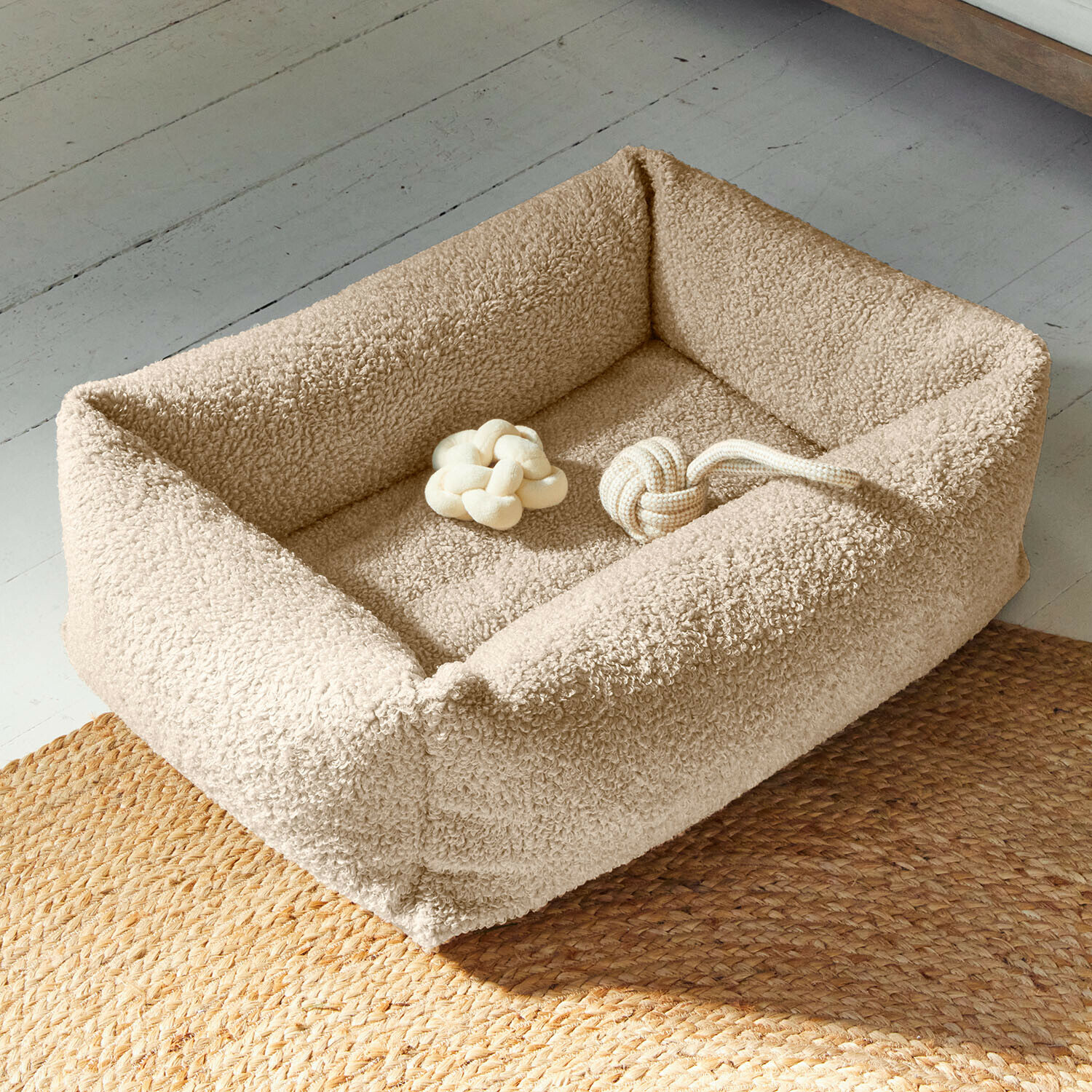 Letto per cani Giolena Cuccia per cane beige in materiale morbido con due giocattoli a nodo beige su pavimento con tappeto intrecciato