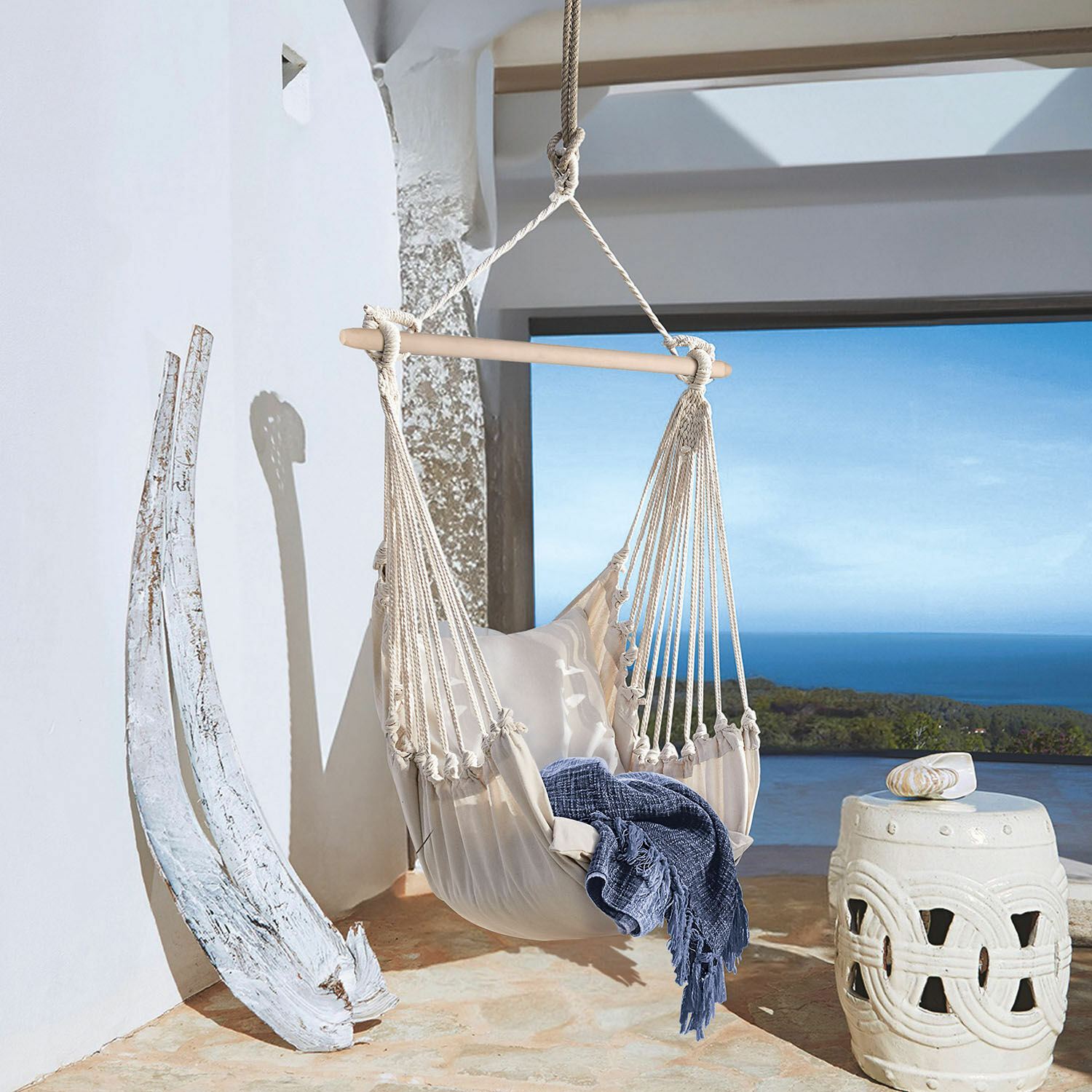 Poltrona sospesa Leano Amaca beige in cotone con barra in legno e coperta blu su terrazza con vista mare