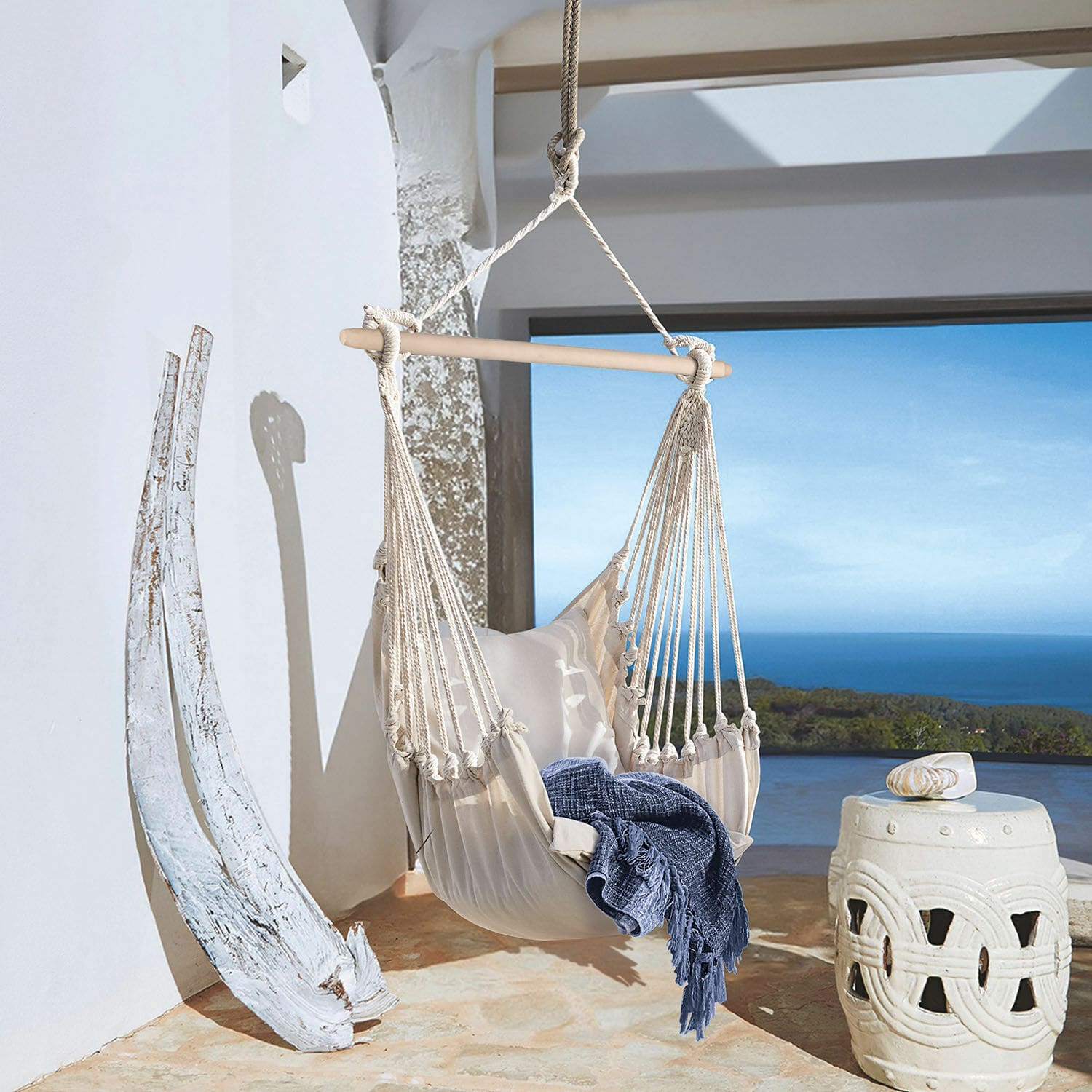 Poltrona sospesa Leano Amaca beige in cotone con barra in legno e coperta blu su terrazza con vista mare