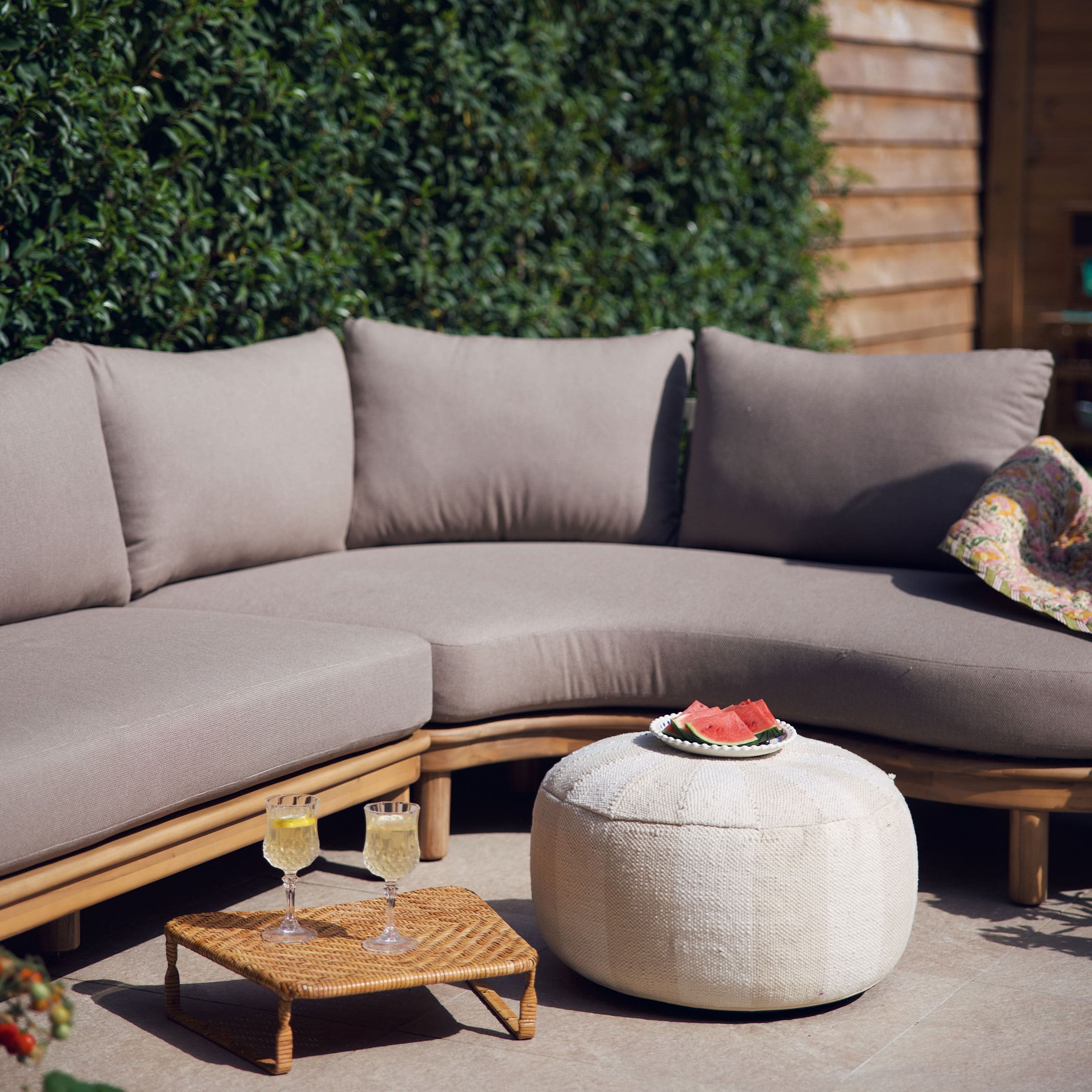 Divano beige con cuscini su struttura in legno e pouf bianco su terrazza con decorazioni estive