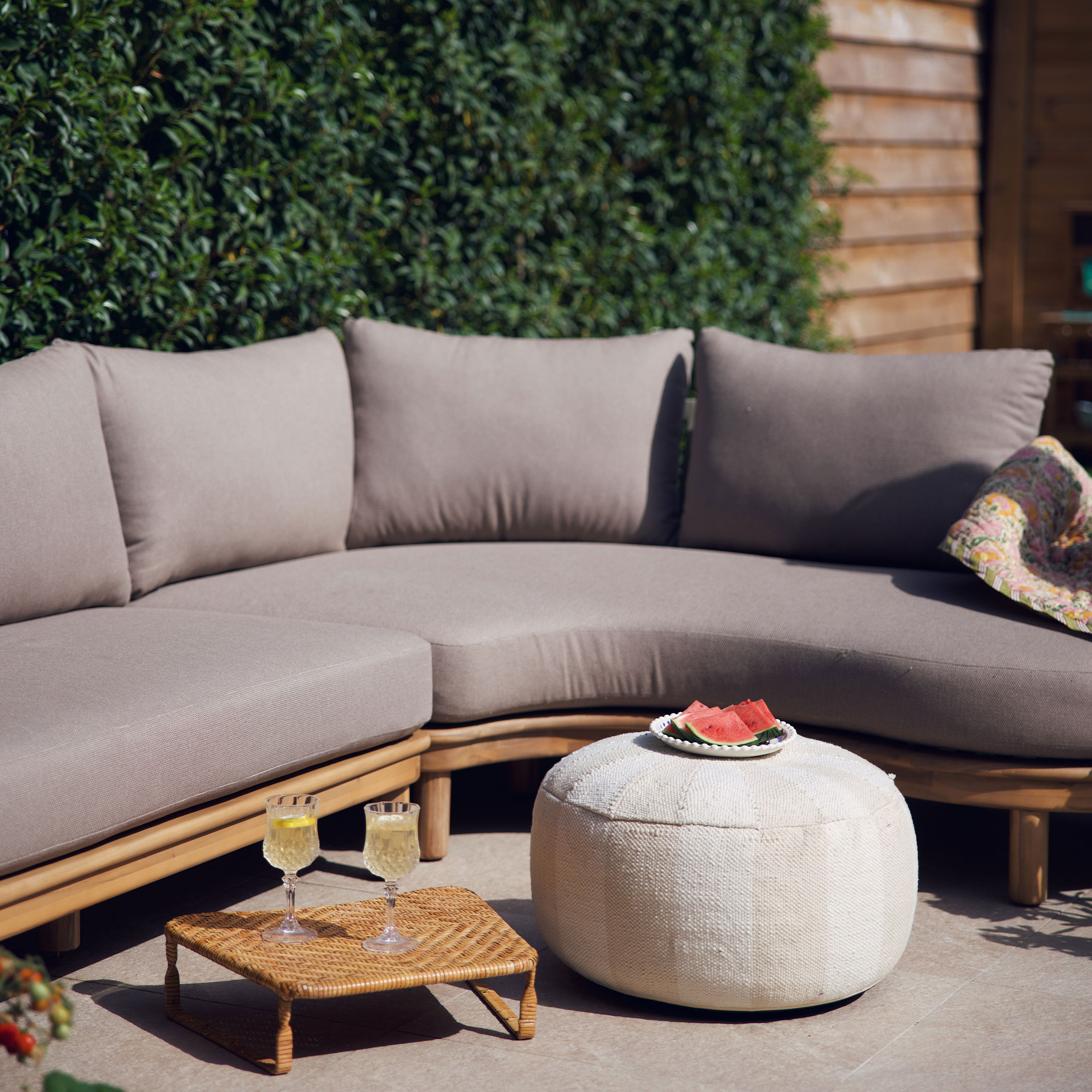 Divano beige con cuscini su struttura in legno e pouf bianco su terrazza con decorazioni estive