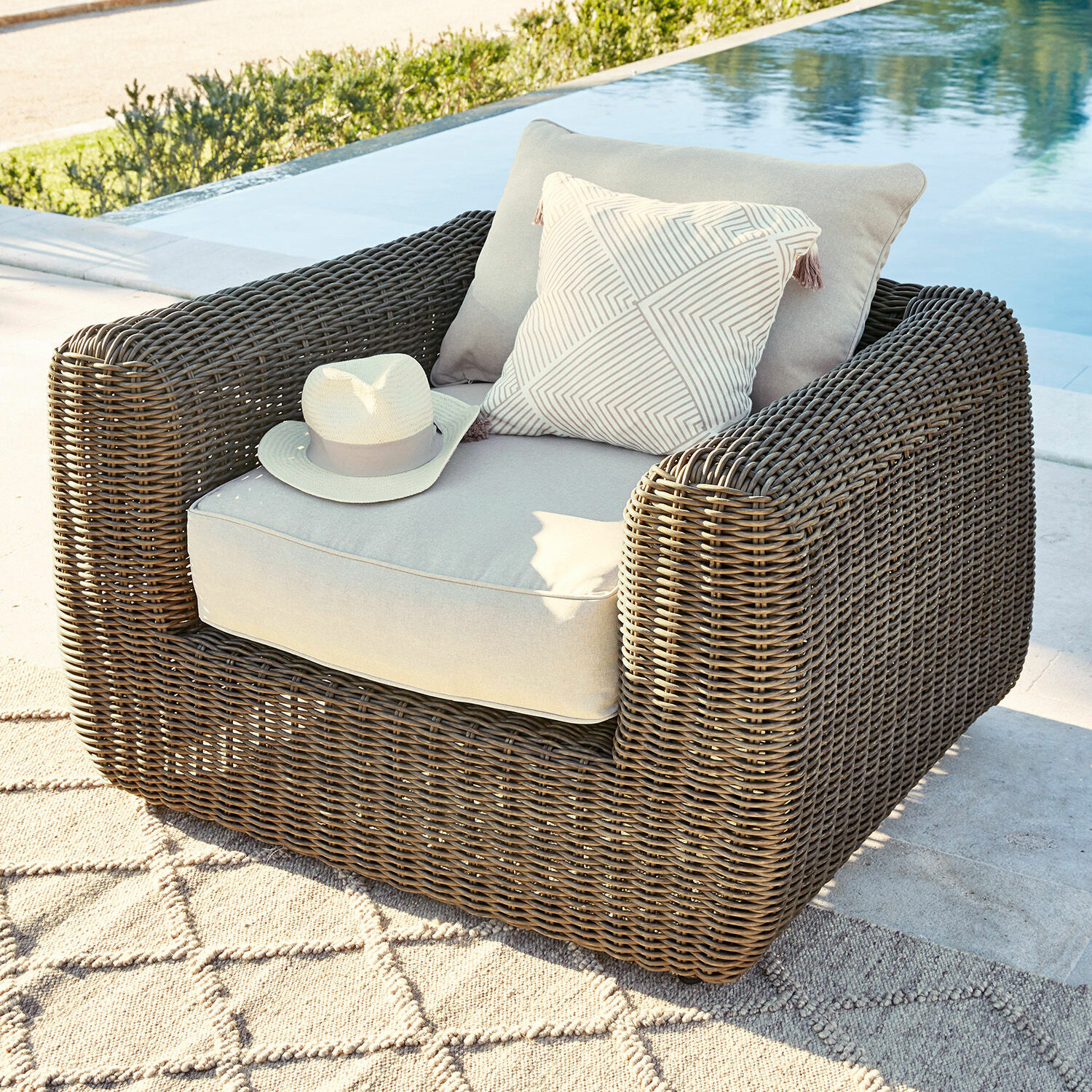 Poltrona Bogdanne Poltrona da giardino in rattan marrone con cuscini grigio chiaro e cuscino decorativo vicino alla piscina