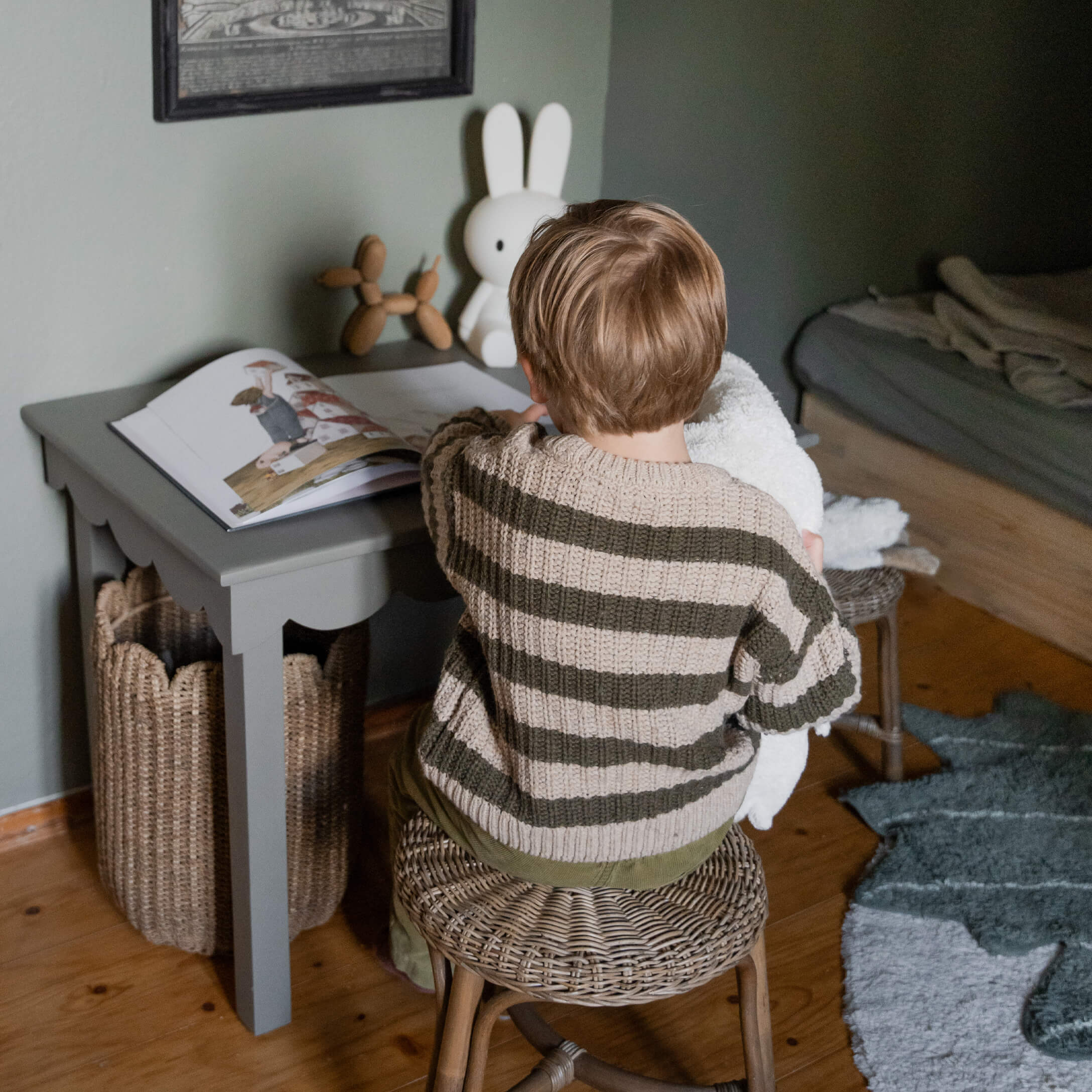 Bambino con maglione a righe seduto su sgabello in vimini a tavolo grigio con libro e lampada coniglio