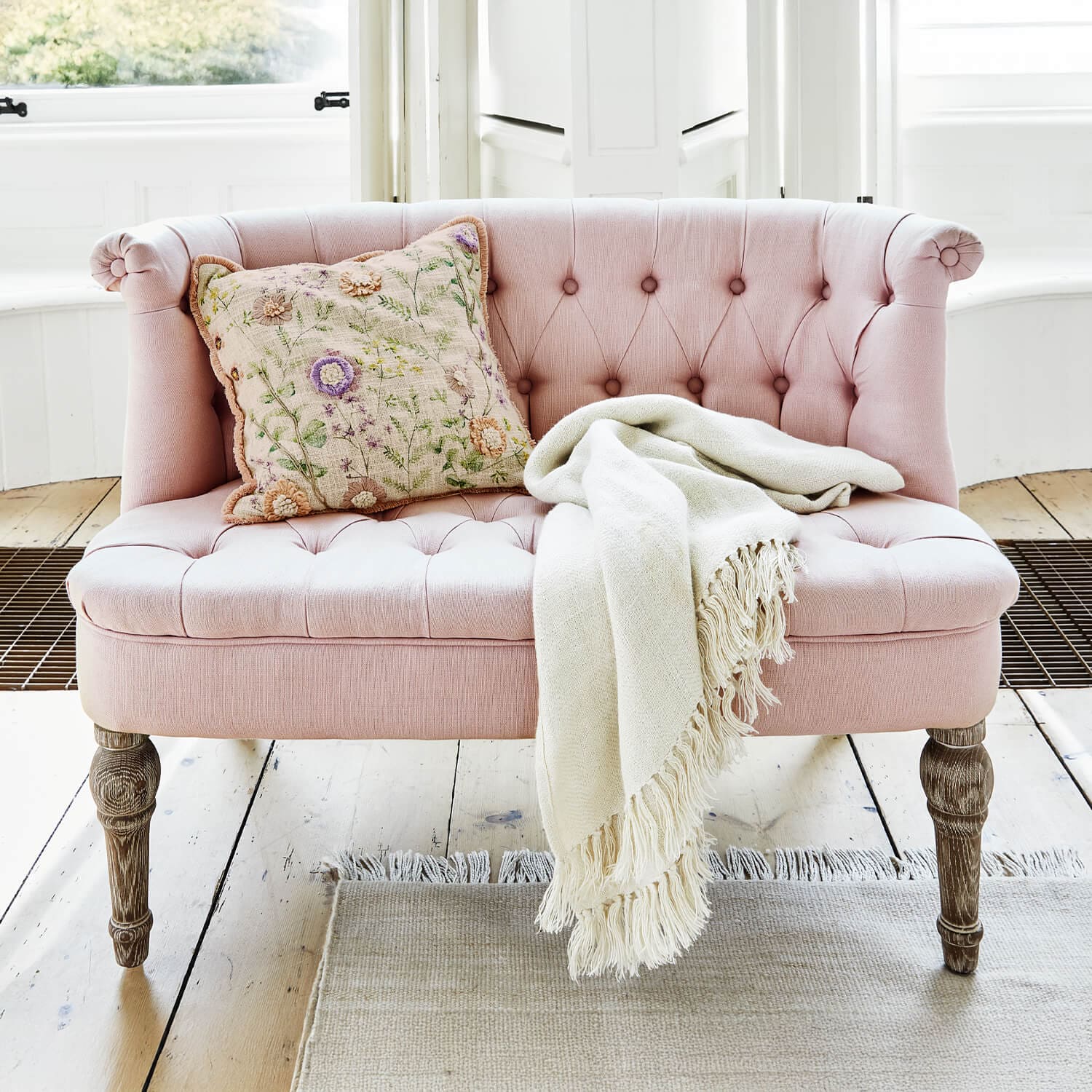 Divano imbottito rosa con gambe in legno, cuscino decorativo e coperta crema su pavimento chiaro