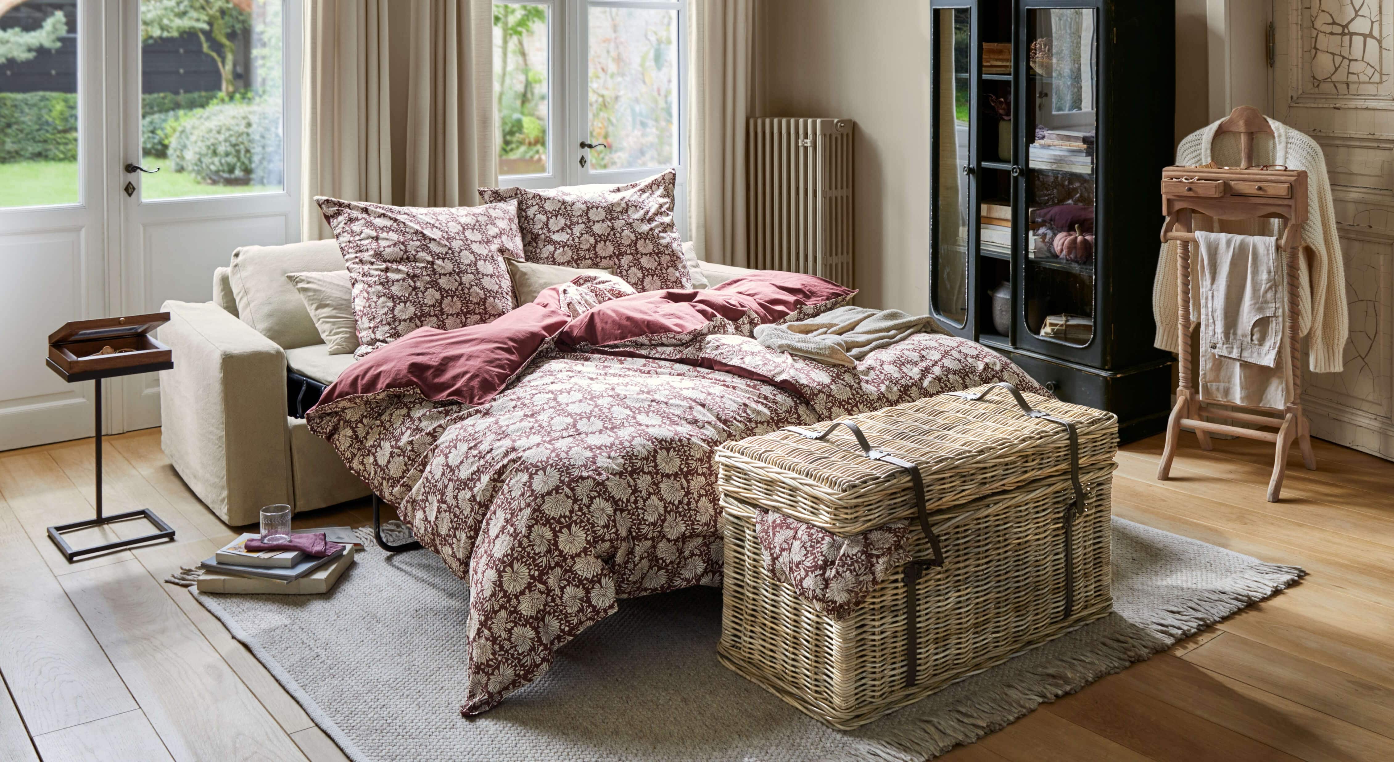 Divano letto beige con biancheria da letto bordeaux a motivi floreali in soggiorno luminoso con pavimento in legno