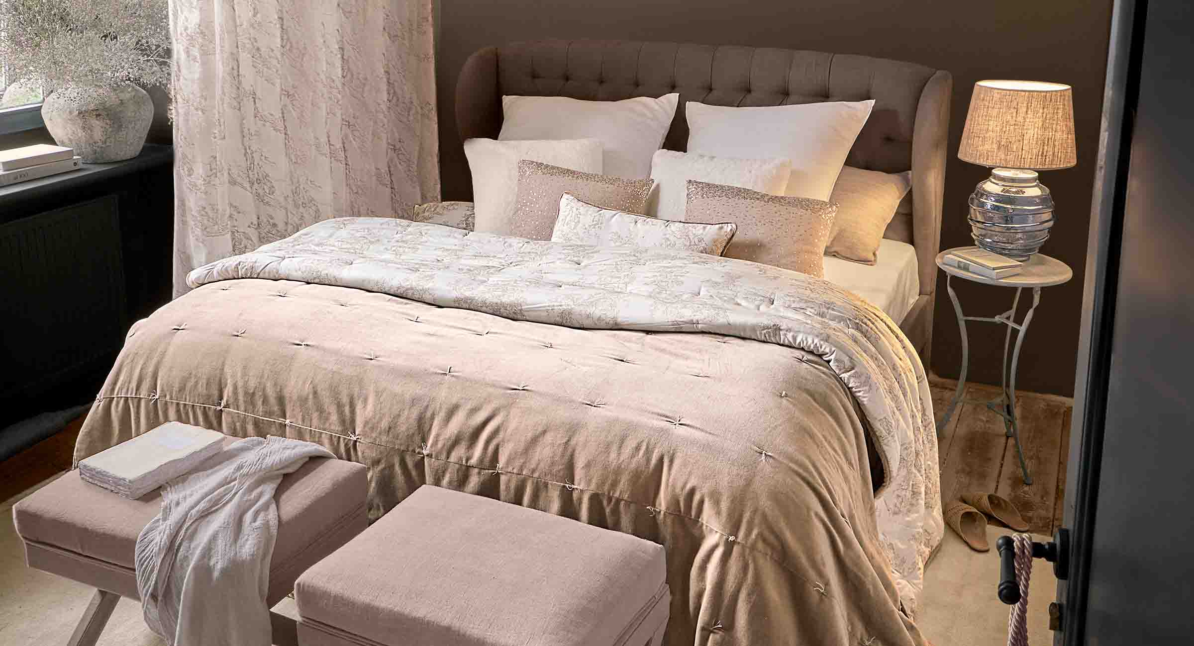Camera da letto accogliente con coperta beige e cuscini su letto imbottito, lampada su comodino