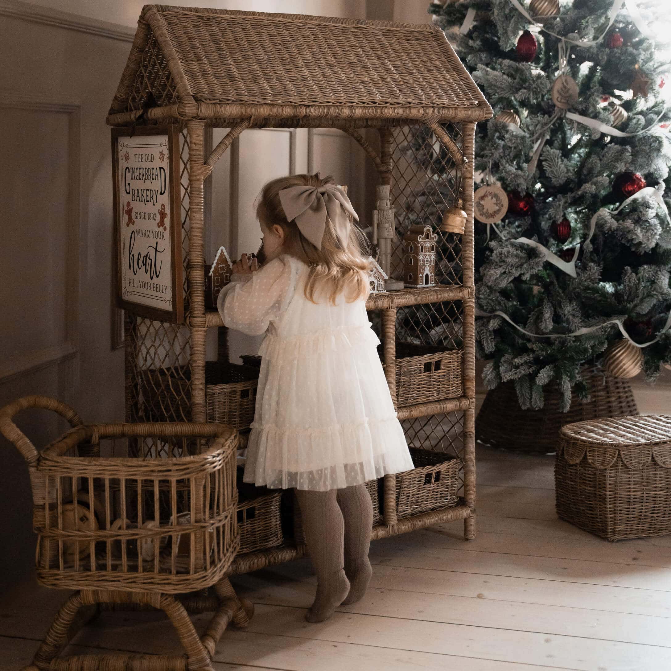 Bambina in vestito bianco davanti a scaffale in vimini con casette di pan di zenzero vicino all'albero di Natale