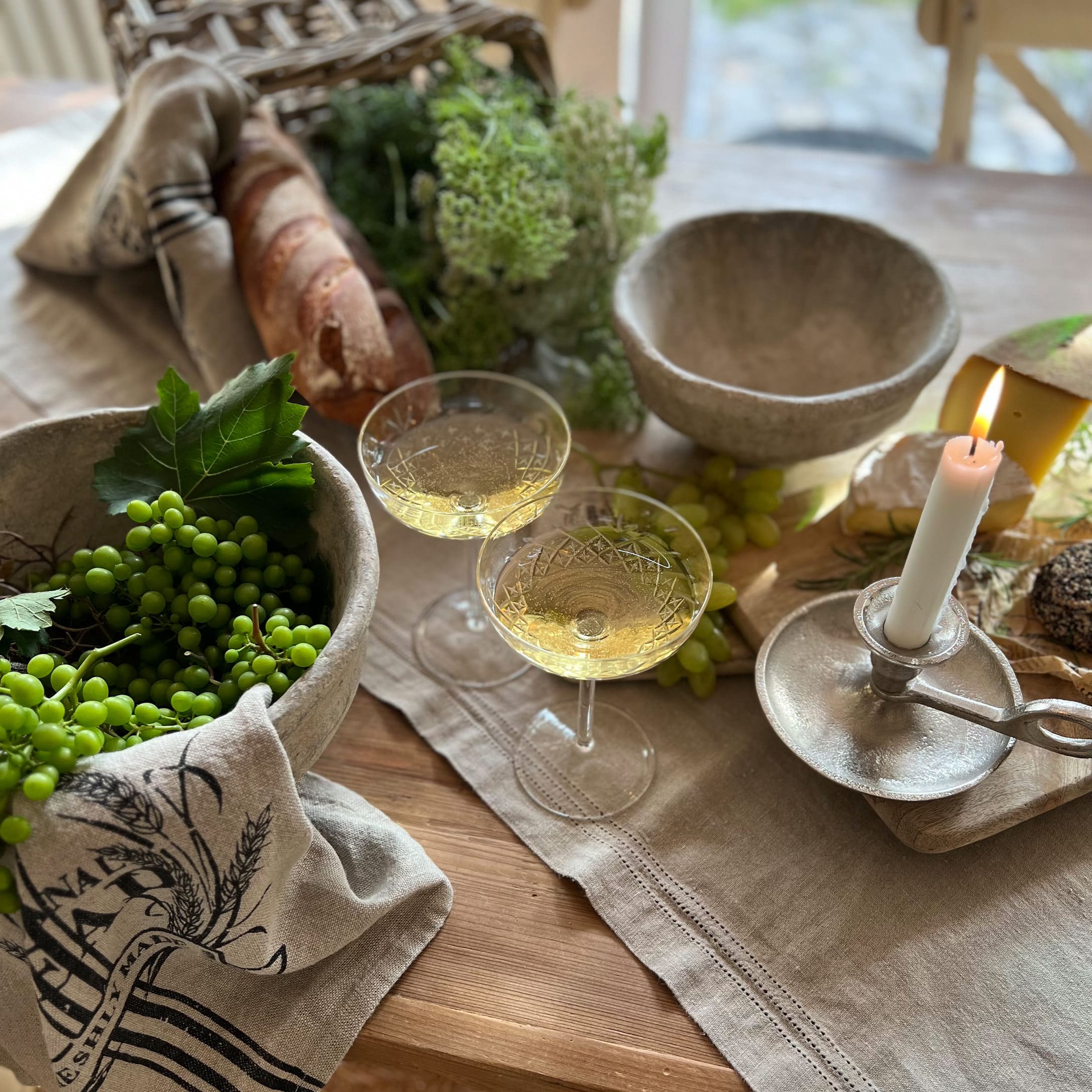 Tavolo rustico con ciotole in pietra, candela accesa, bicchieri di vino bianco, pane e uva verde