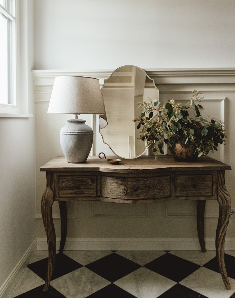 Scrivania in legno con specchio, lampada e decorazione vegetale su pavimento bianco e nero in stanza luminosa