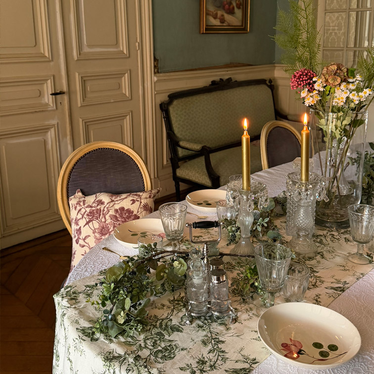 Tavolo elegante con tovaglia bianca a motivi verdi, candele accese e decorazioni floreali in interno classico