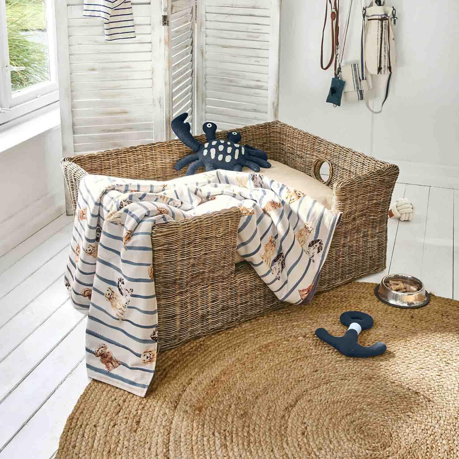 Cuccia in vimini per cane con cuscino beige, coperta a righe con motivo cani, giochi e ciotola