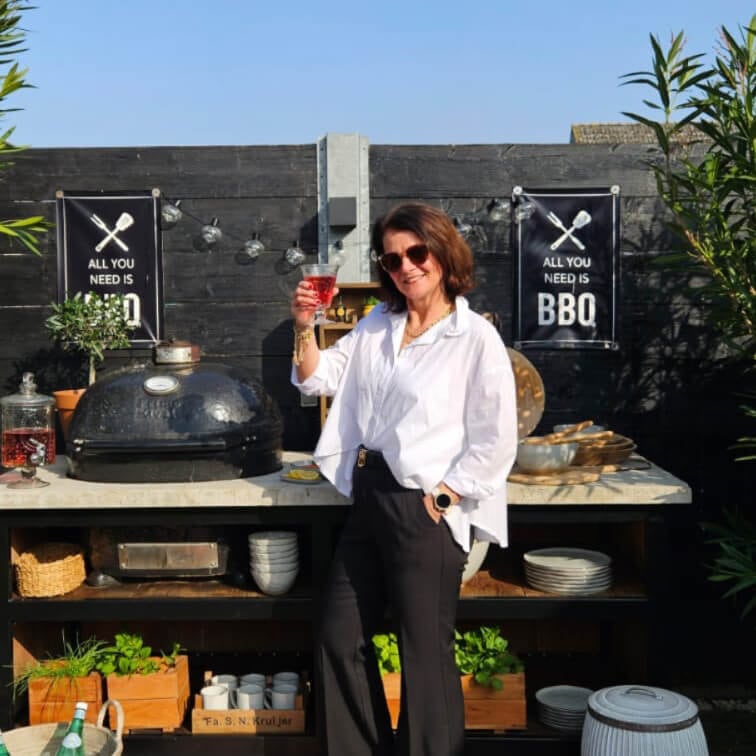 Donna con camicia bianca e pantaloni neri accanto a barbecue esterno con insegna BBQ su muro nero