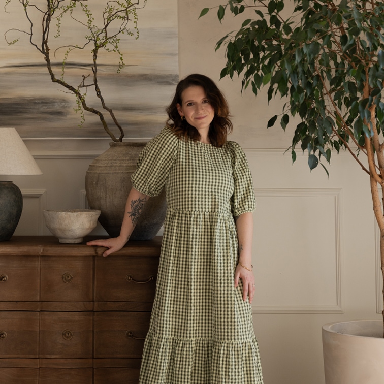 Donna con vestito verde a quadri vicino a cassettiera in legno in ambiente accogliente con piante