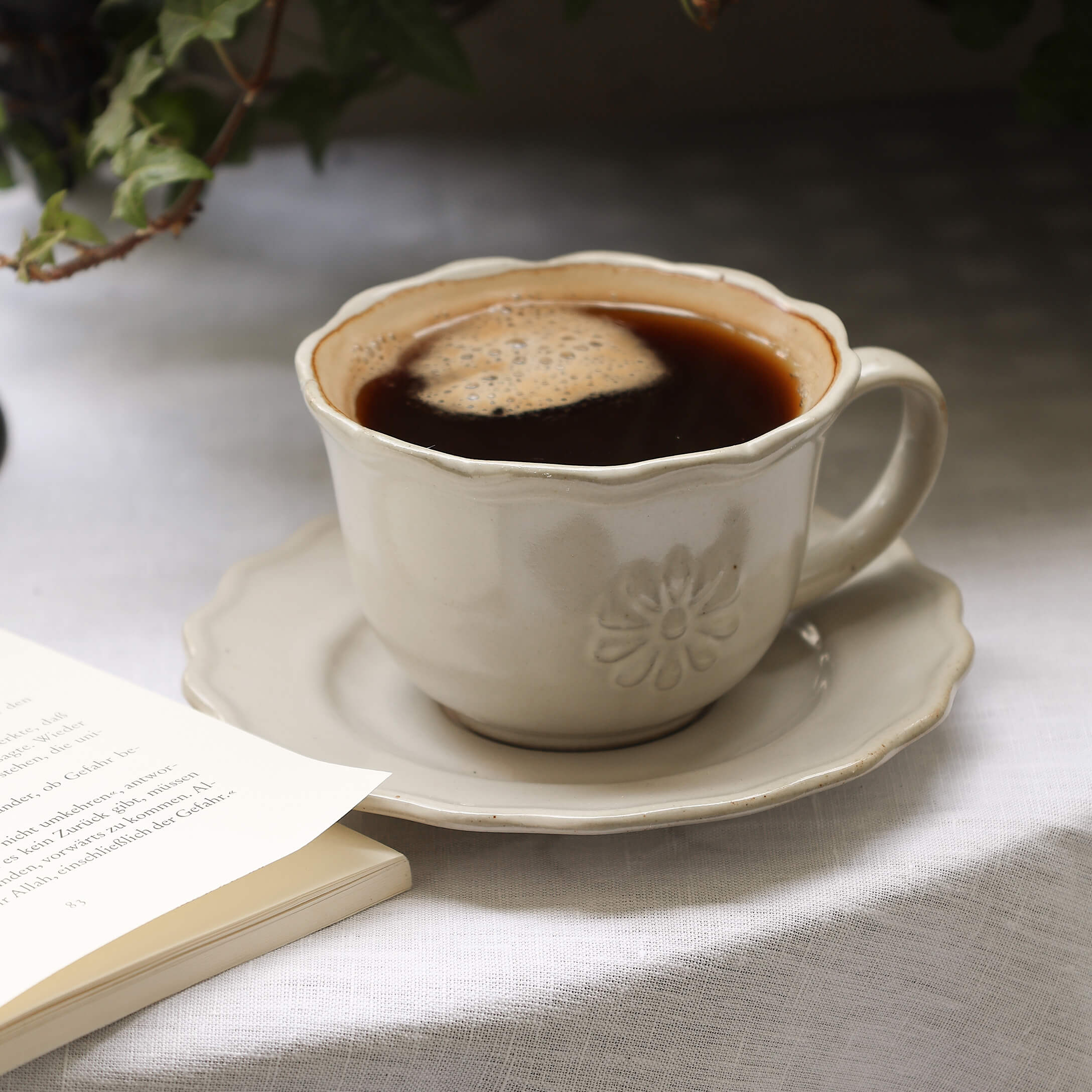 Tazza in ceramica beige con caffè su piattino su tovaglia bianca accanto a libro aperto