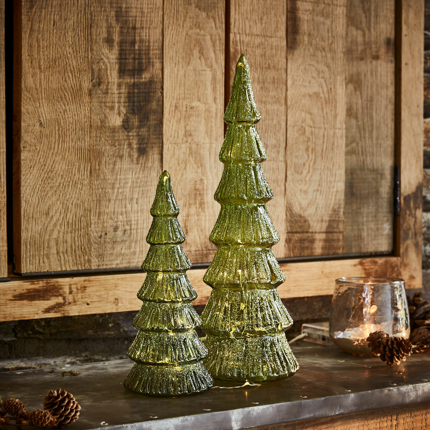 Coppia di alberi di Natale a LED Merrton Due decorazioni natalizie a forma di albero in vetro verde con luci LED su sfondo in legno, addobbi festivi