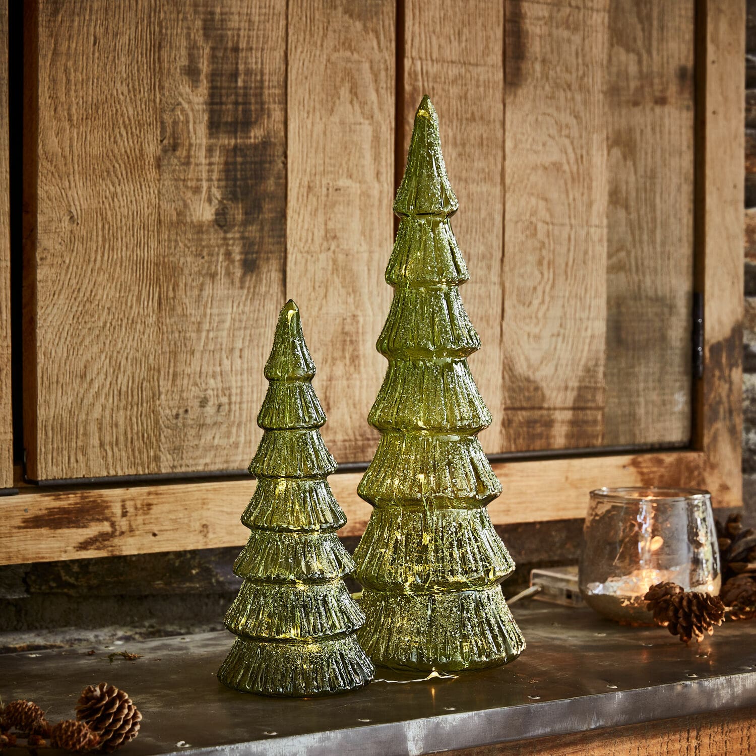 Coppia di alberi di Natale a LED Merrton Due decorazioni natalizie a forma di albero in vetro verde con luci LED su sfondo in legno, addobbi festivi