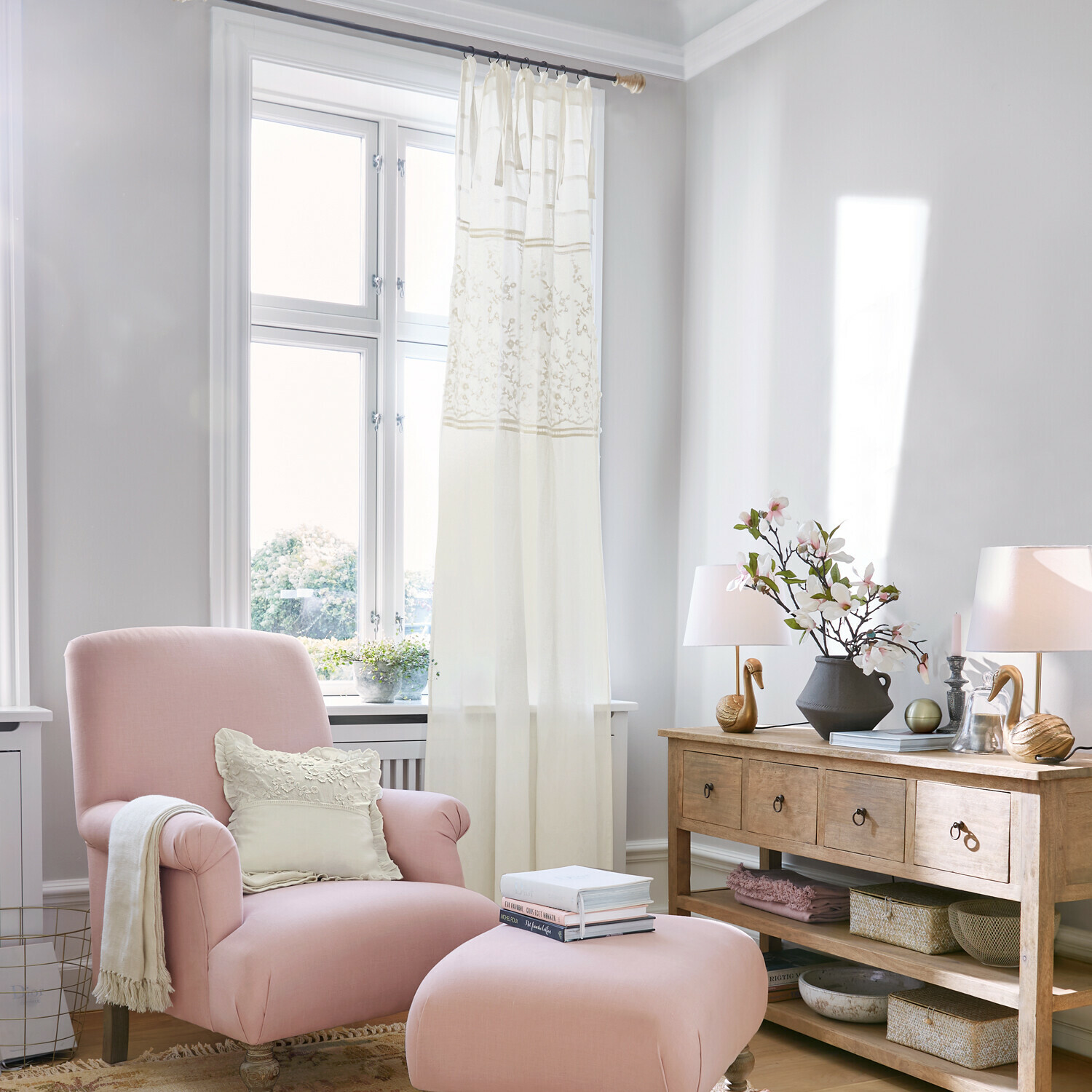 Tenda Lunérie Poltrona rosa con poggiapiedi e cuscino crema in soggiorno luminoso con comò in legno e decorazioni