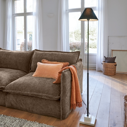 Divano marrone in tessuto morbido con cuscino e coperta arancione, lampada da terra nera con base in marmo in soggiorno luminoso
