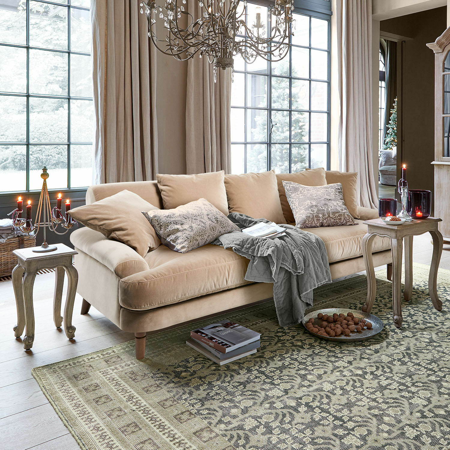 Divano Rivermere Divano beige in velluto con cuscini e coperta grigia in soggiorno luminoso con tappeto e candele