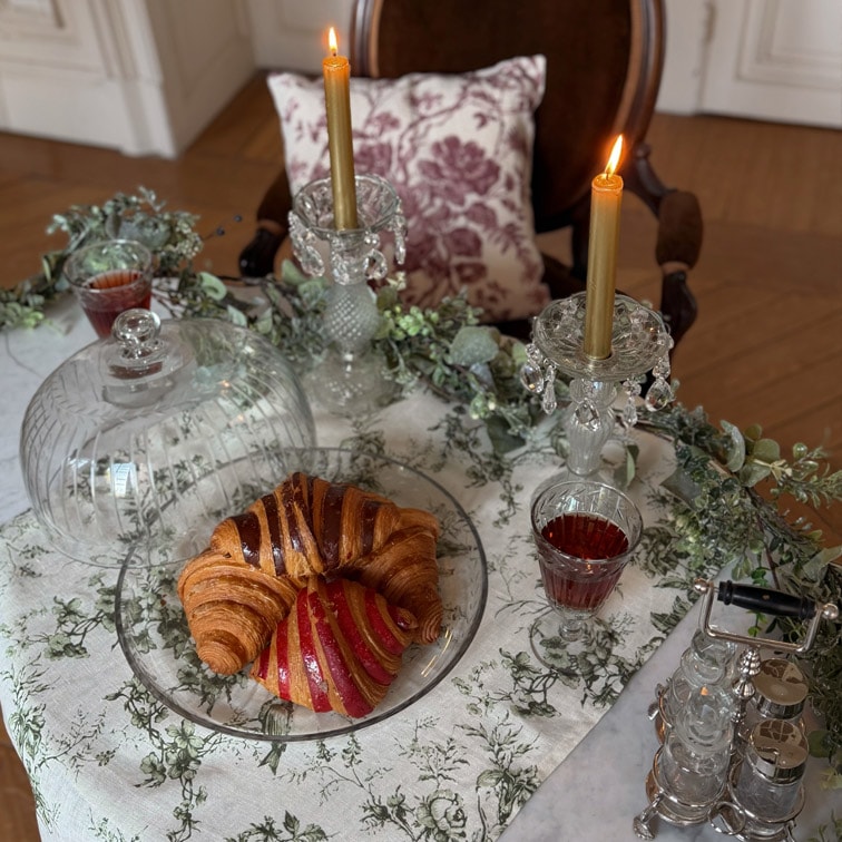 Piatto di vetro con croissant e dolce su tavolo con tovaglia bianca, due candele dorate in candelabri di cristallo