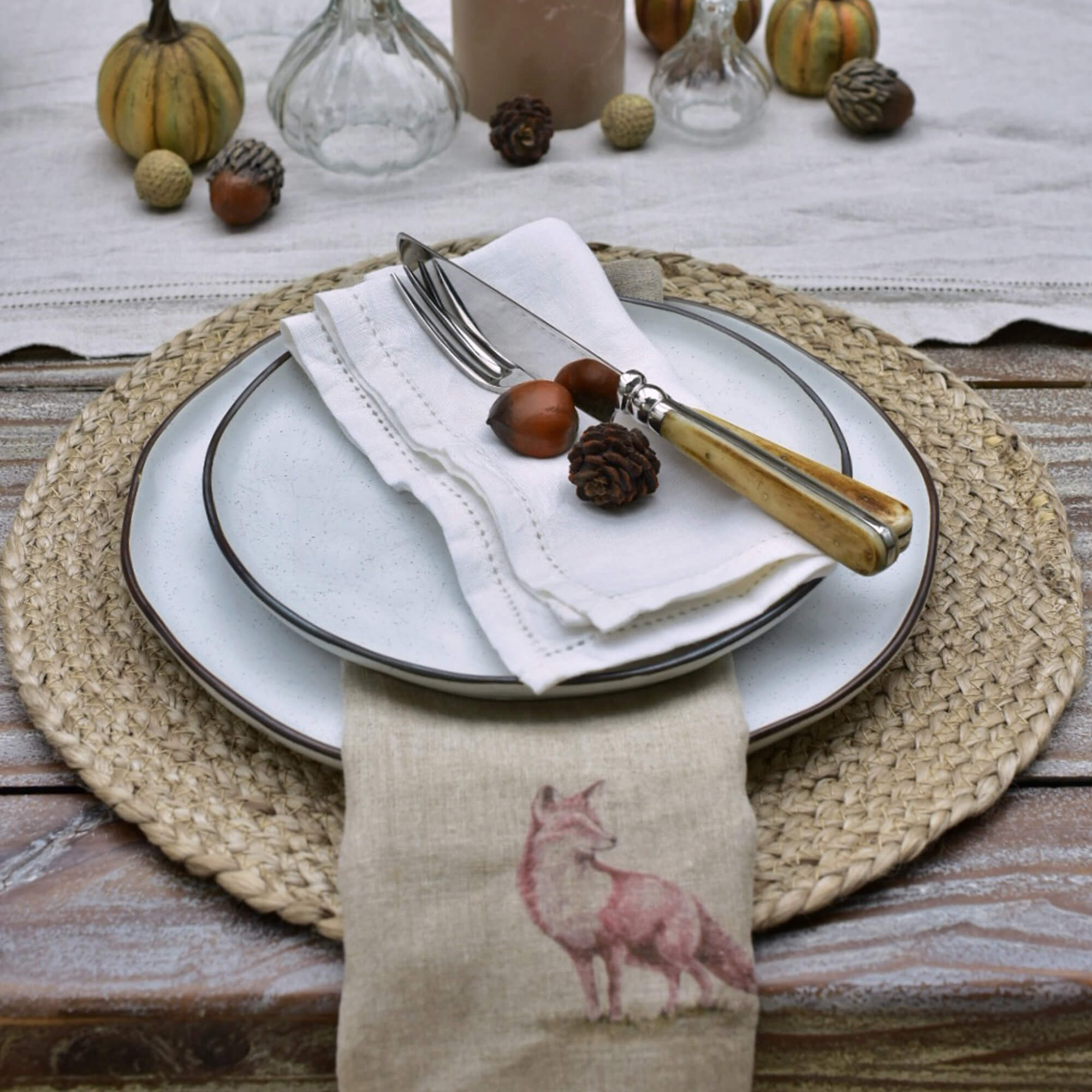 Servizio tavola con piatti in ceramica bianca, posate con manico in legno chiaro e tovagliolo di lino con stampa volpe