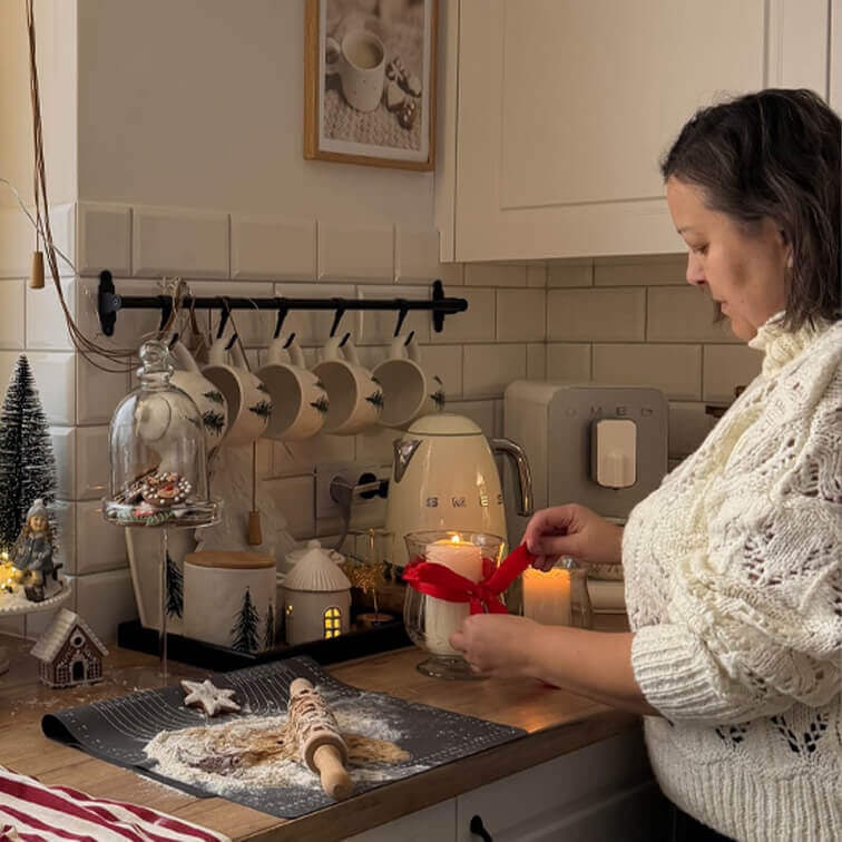 Donna con maglione bianco decora candela con nastro rosso in cucina con piano in legno e decorazioni natalizie