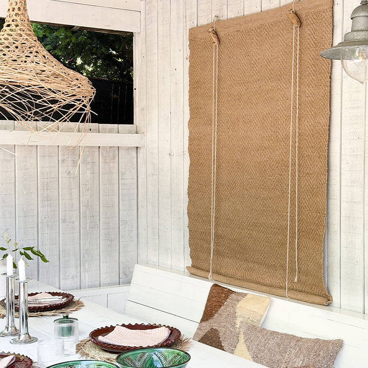 Parete in legno bianco con tenda a rullo in juta naturale e tavolo decorato con cuscini in stile boho
