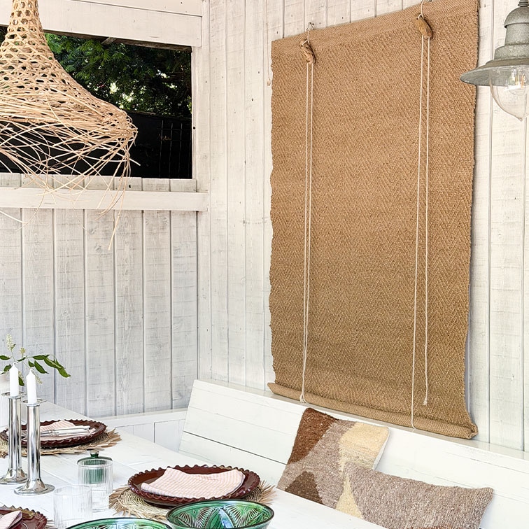 Parete in legno bianco con tenda a rullo in juta naturale e tavolo decorato con cuscini in stile boho