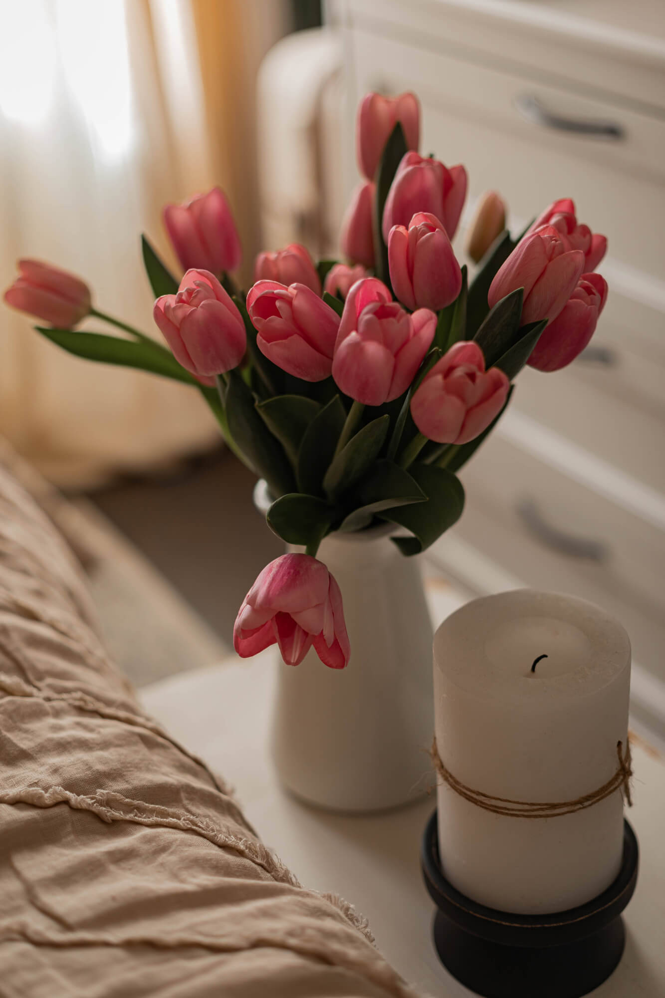 Mazzo di tulipani rosa in brocca bianca accanto a candela bianca su comodino in camera