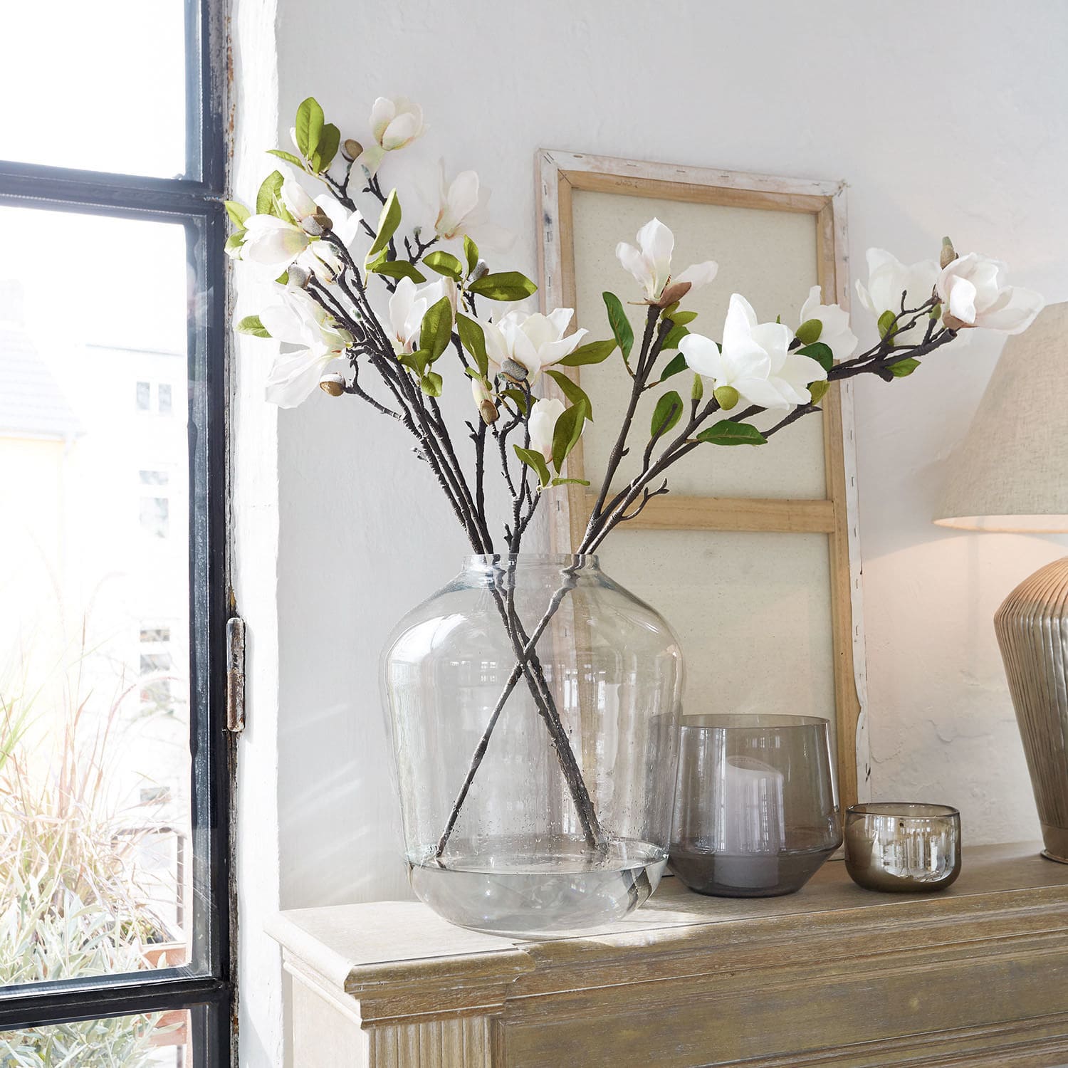 Coppia di fiori decorativi Mariella Vaso in vetro trasparente con fiori bianchi di magnolia e foglie verdi su mobile in legno