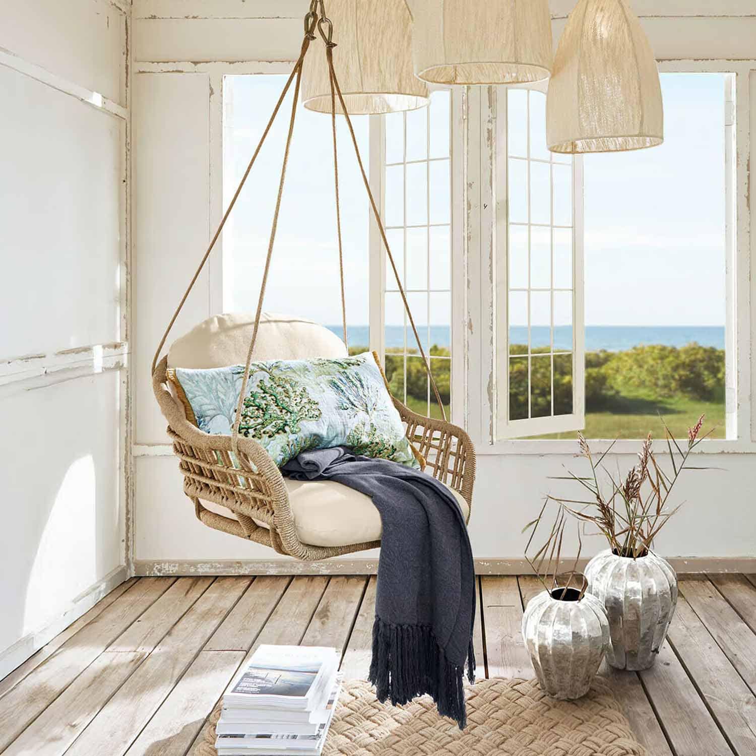 Sedia sospesa in rattan con cuscino crema e coperta blu in ambiente luminoso con vista mare