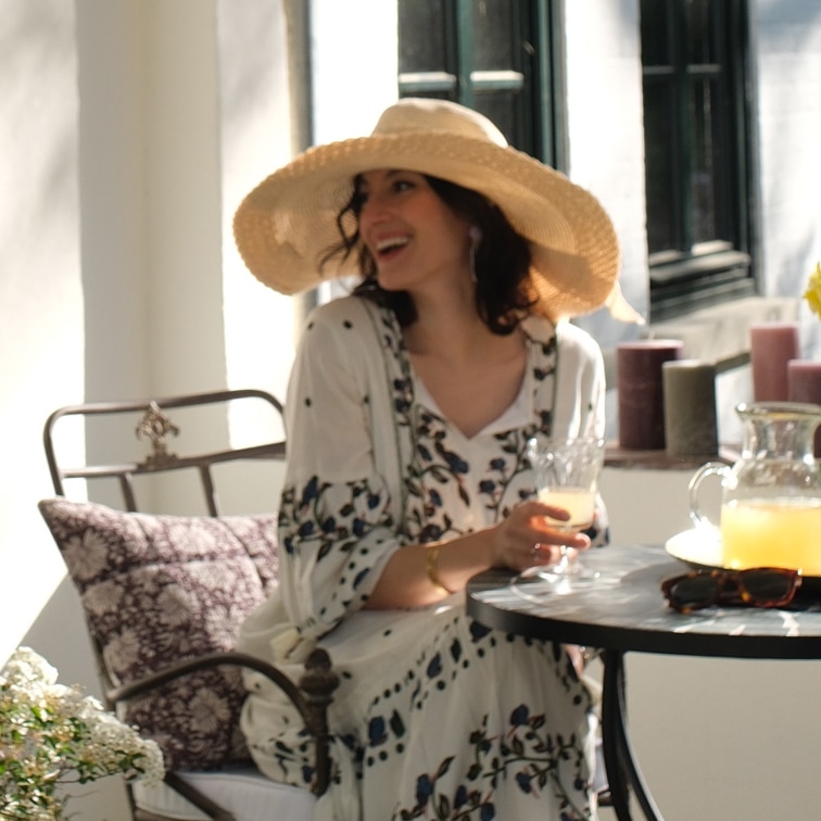 Donna con vestito floreale e cappello di paglia seduta a tavola con bevanda in ambiente luminoso