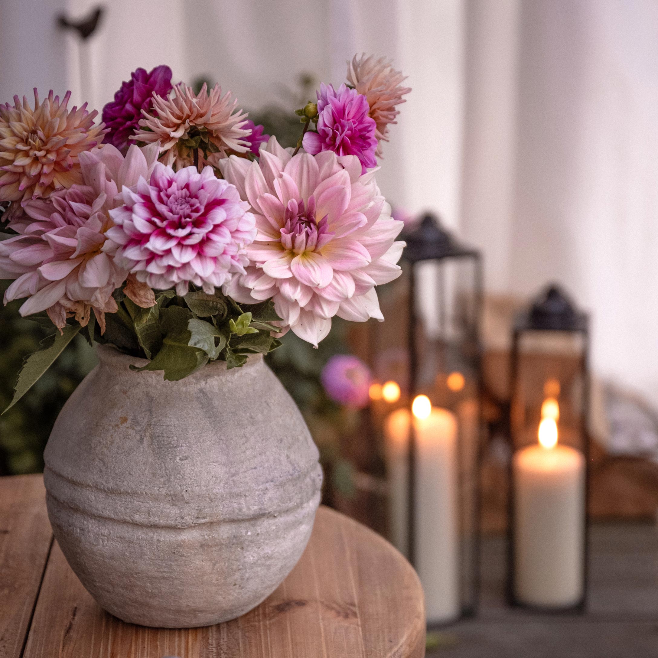 Vaso in ceramica grigia con fiori rosa su tavolo in legno, lanterne e candele accese sullo sfondo