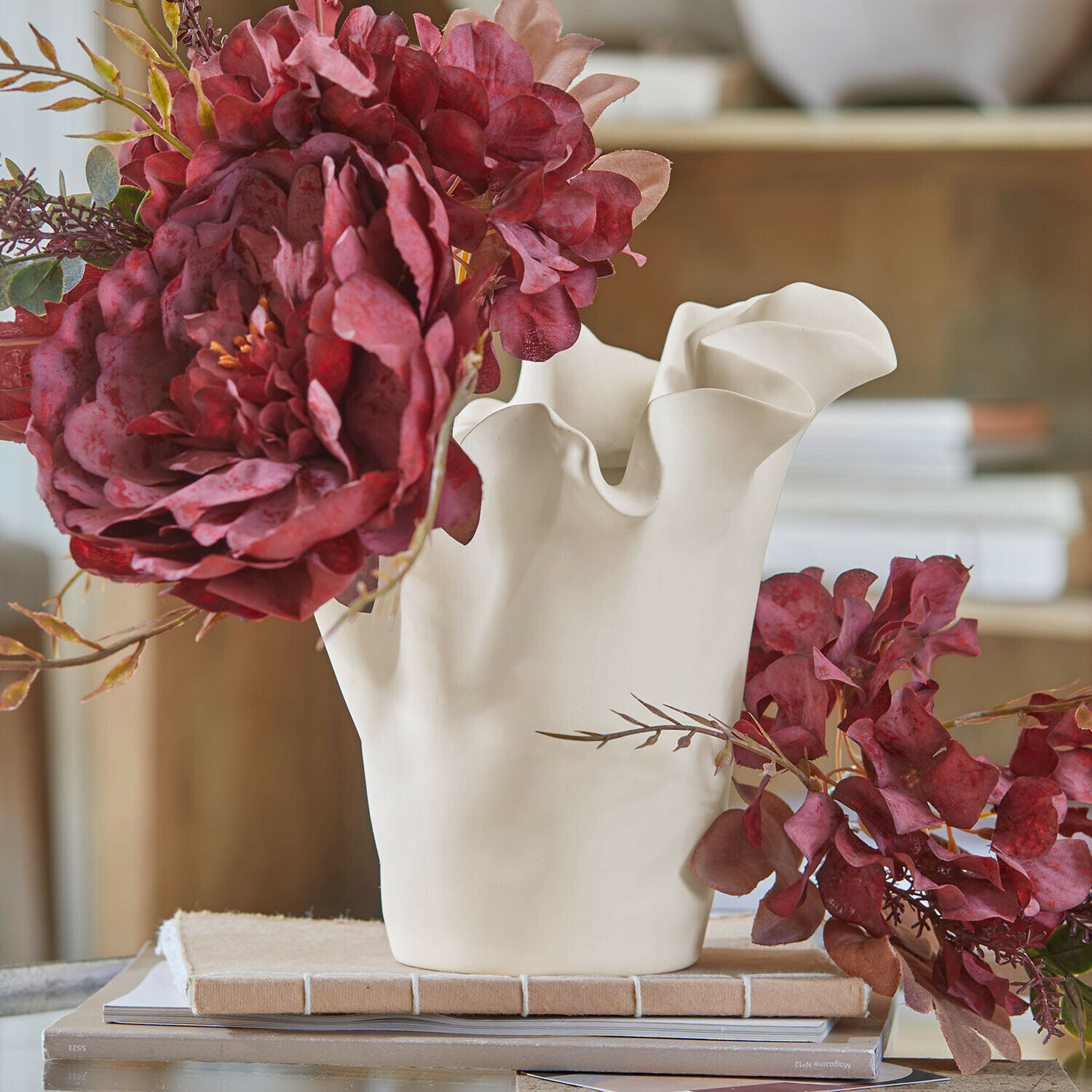 Vaso Elvessa Vaso in ceramica bianca con bordo ondulato e fiori rossi artificiali su tavolo con libri
