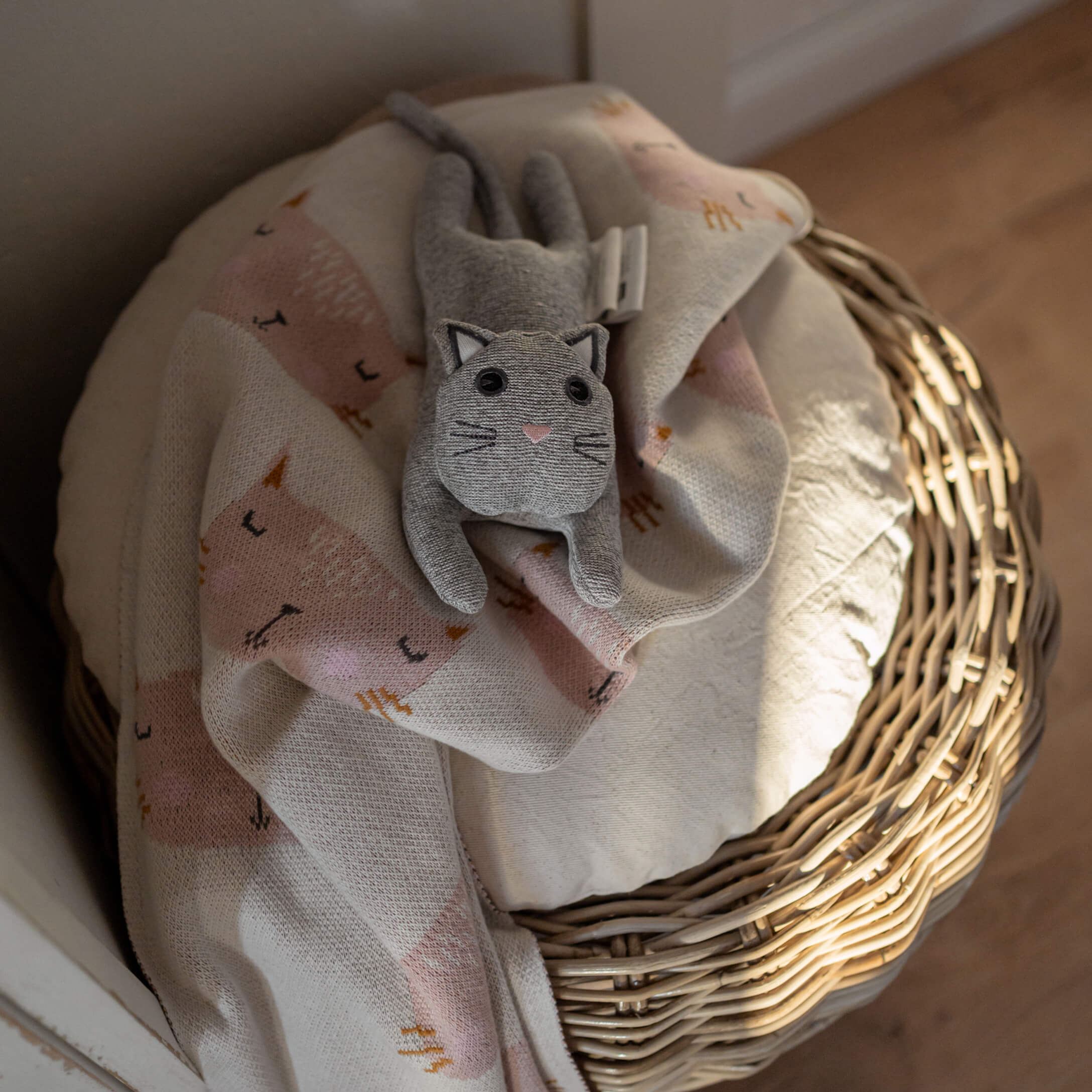 Gatto di peluche grigio su coperta rosa chiaro con motivo gatto su cesta di vimini