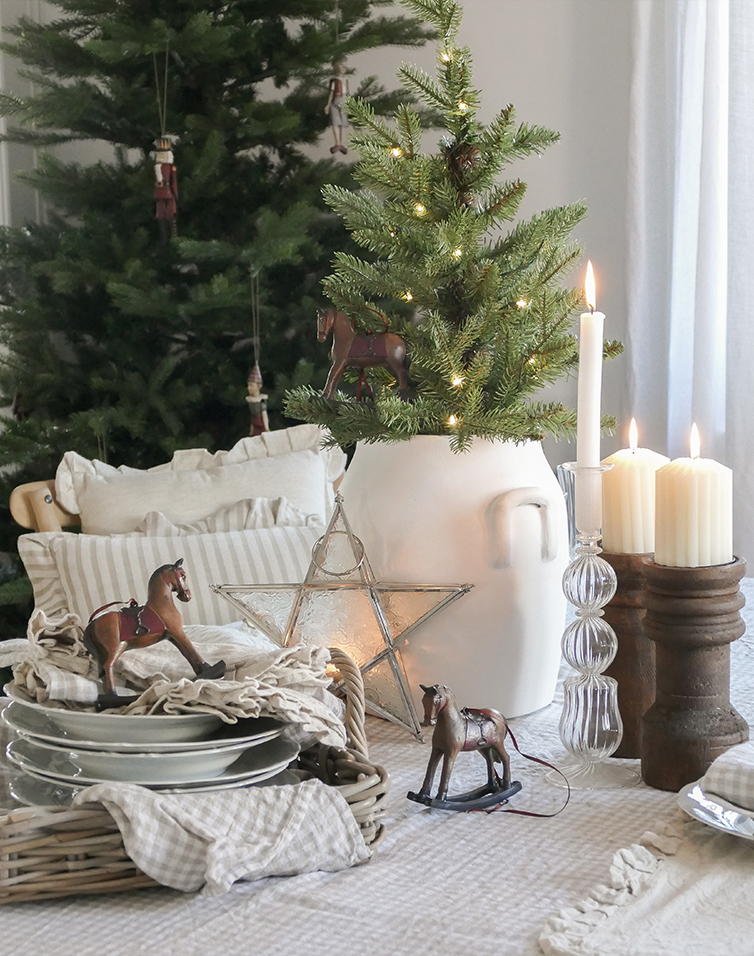 Decorazione natalizia con brocca bianca, albero verde, candele e cavalli a dondolo in legno sul tavolo
